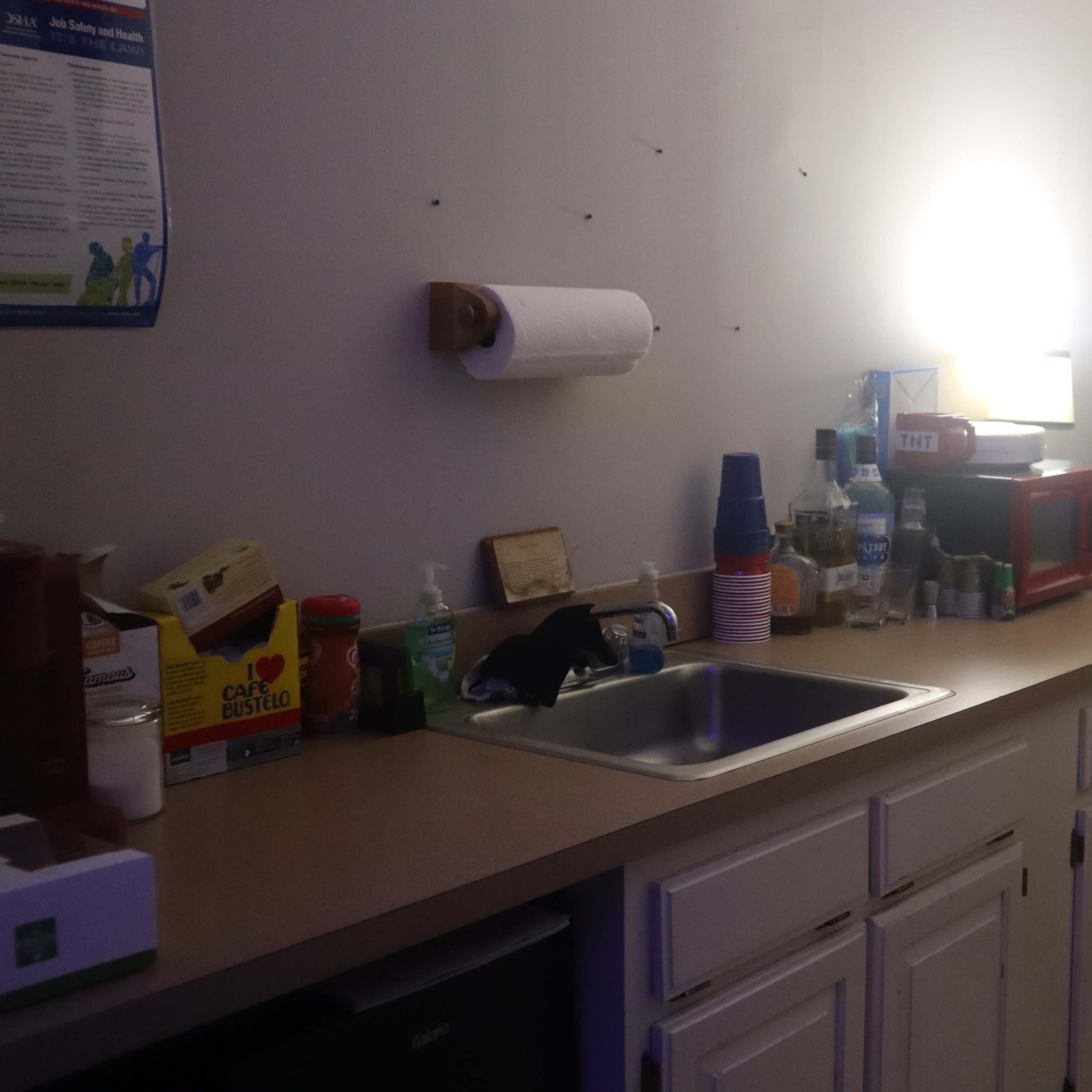 A kitchen counter is lined with various containers and appliances, including a sink and a black refrigerator, under soft overhead lighting.