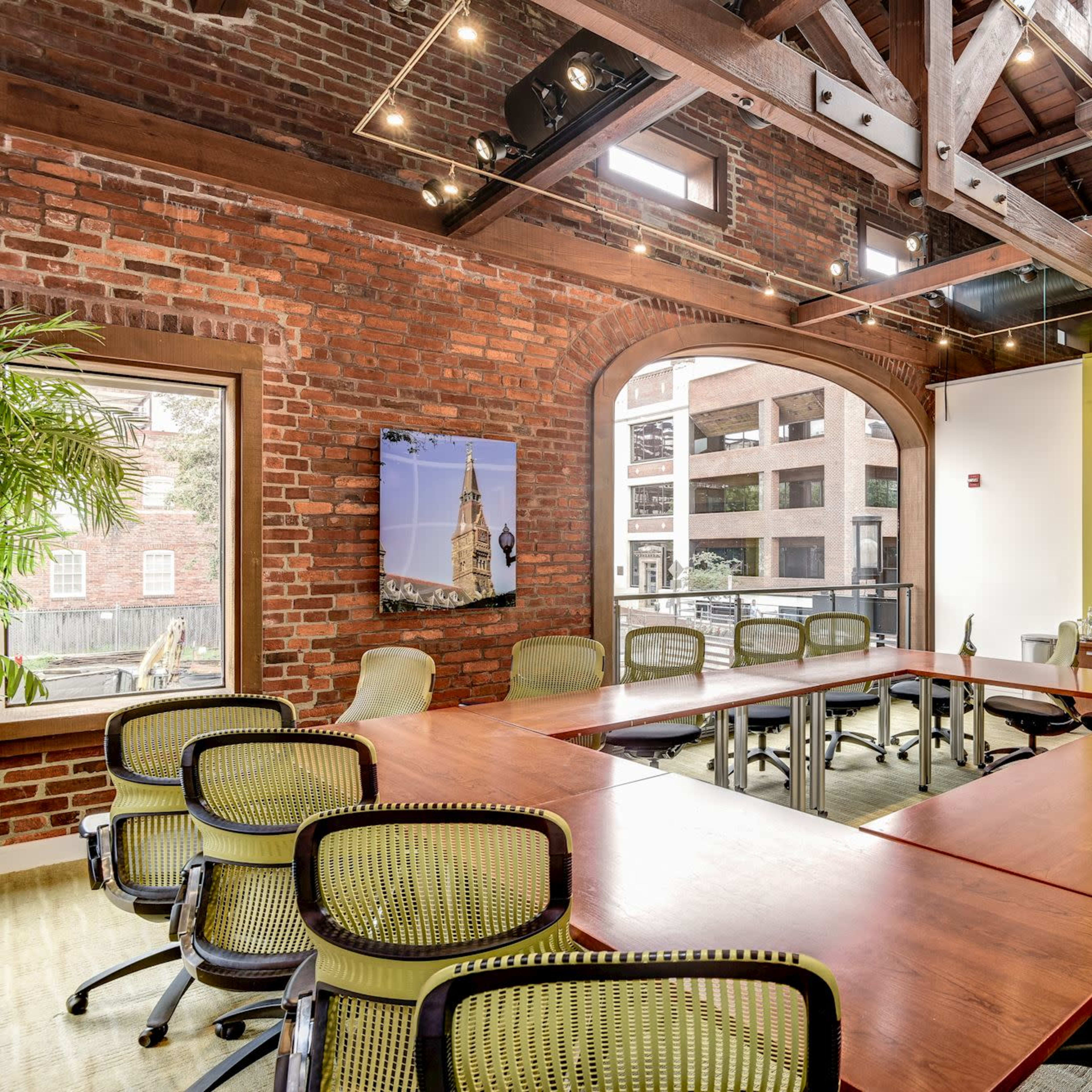 A modern conference room features a long wooden table surrounded by green chairs, with exposed brick walls and large windows allowing natural light.