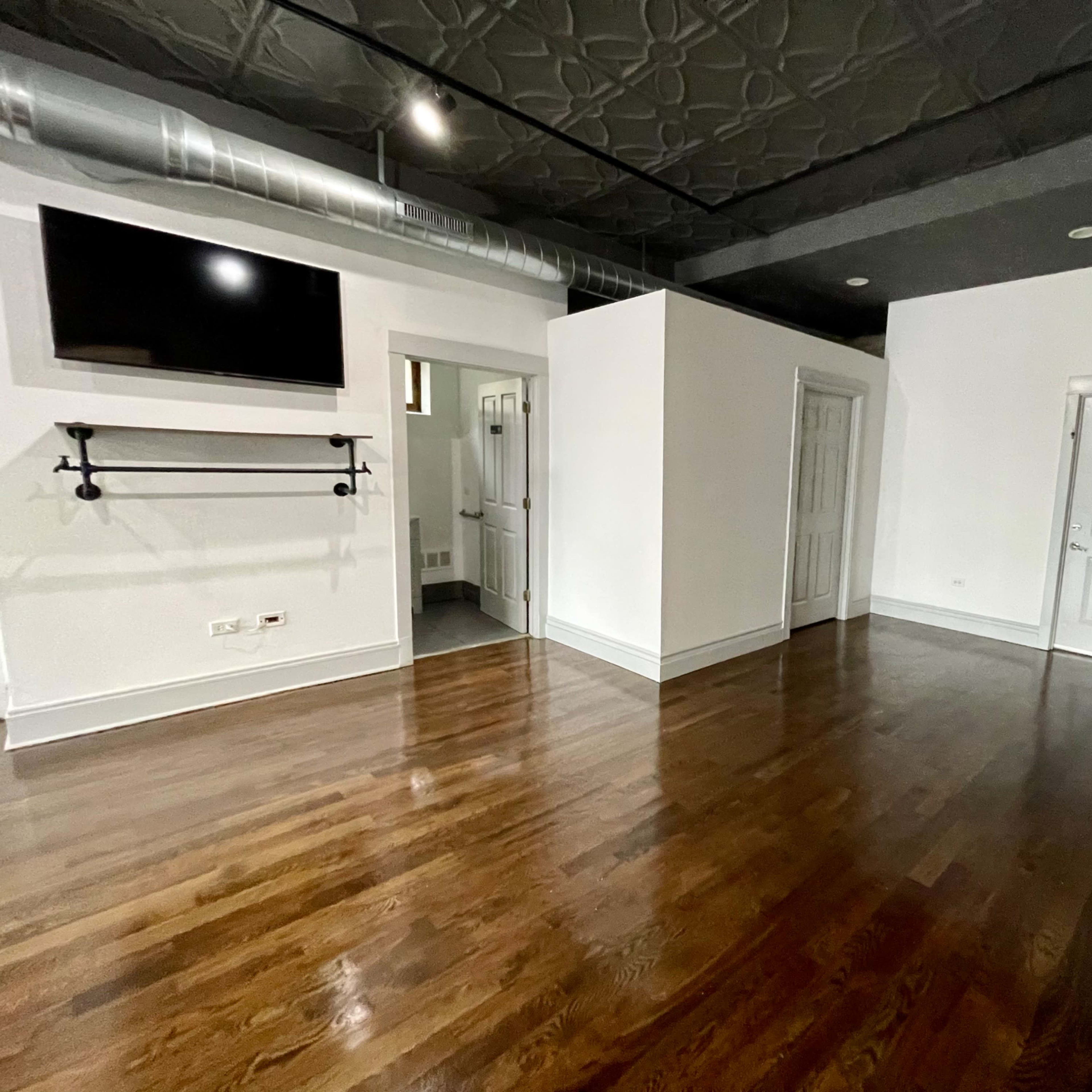 A spacious room with hardwood floors, a flat-screen TV mounted on the wall, and various doorways leading to other areas.