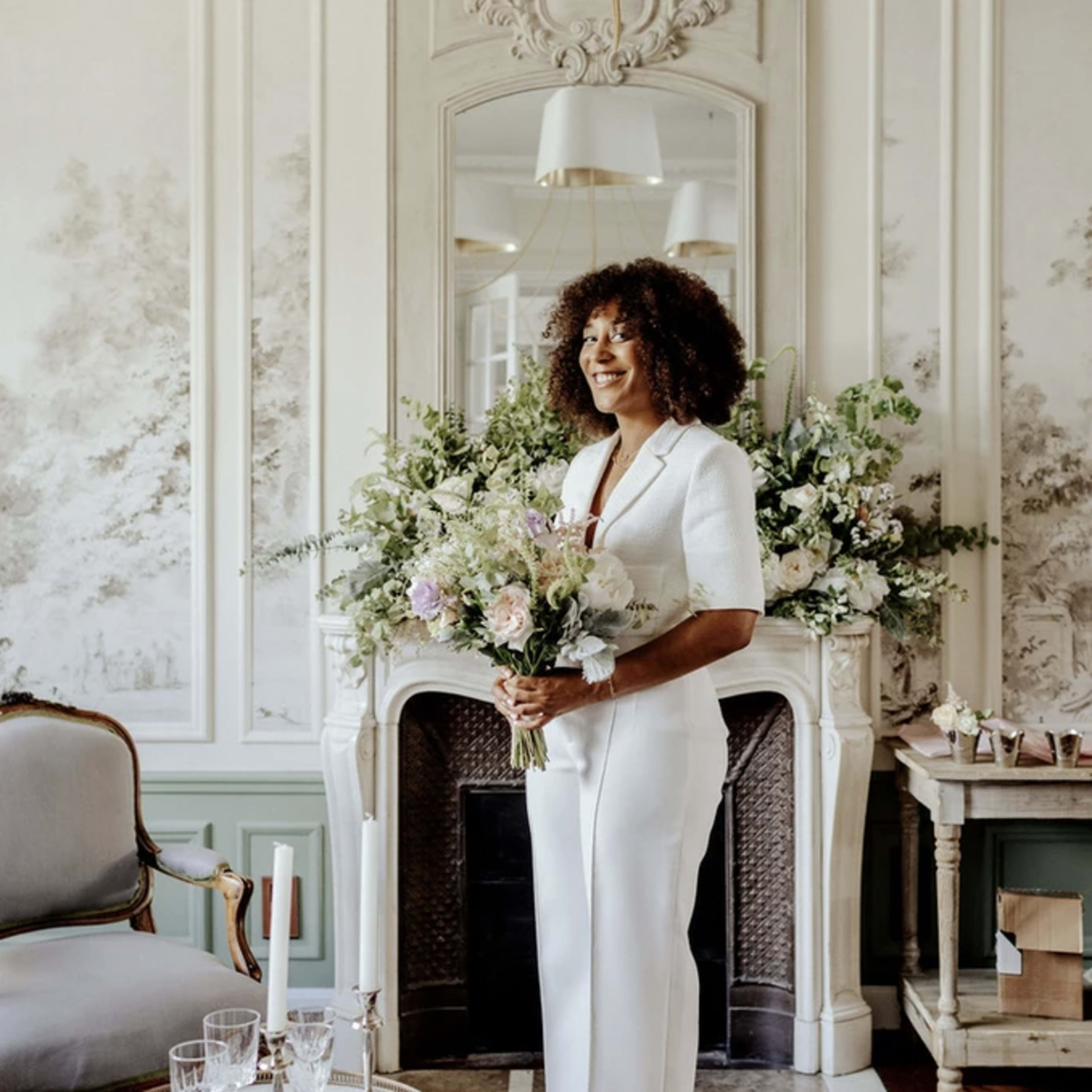 A woman in a white outfit stands beside a decorative fireplace adorned with greenery and flowers in a stylish room.