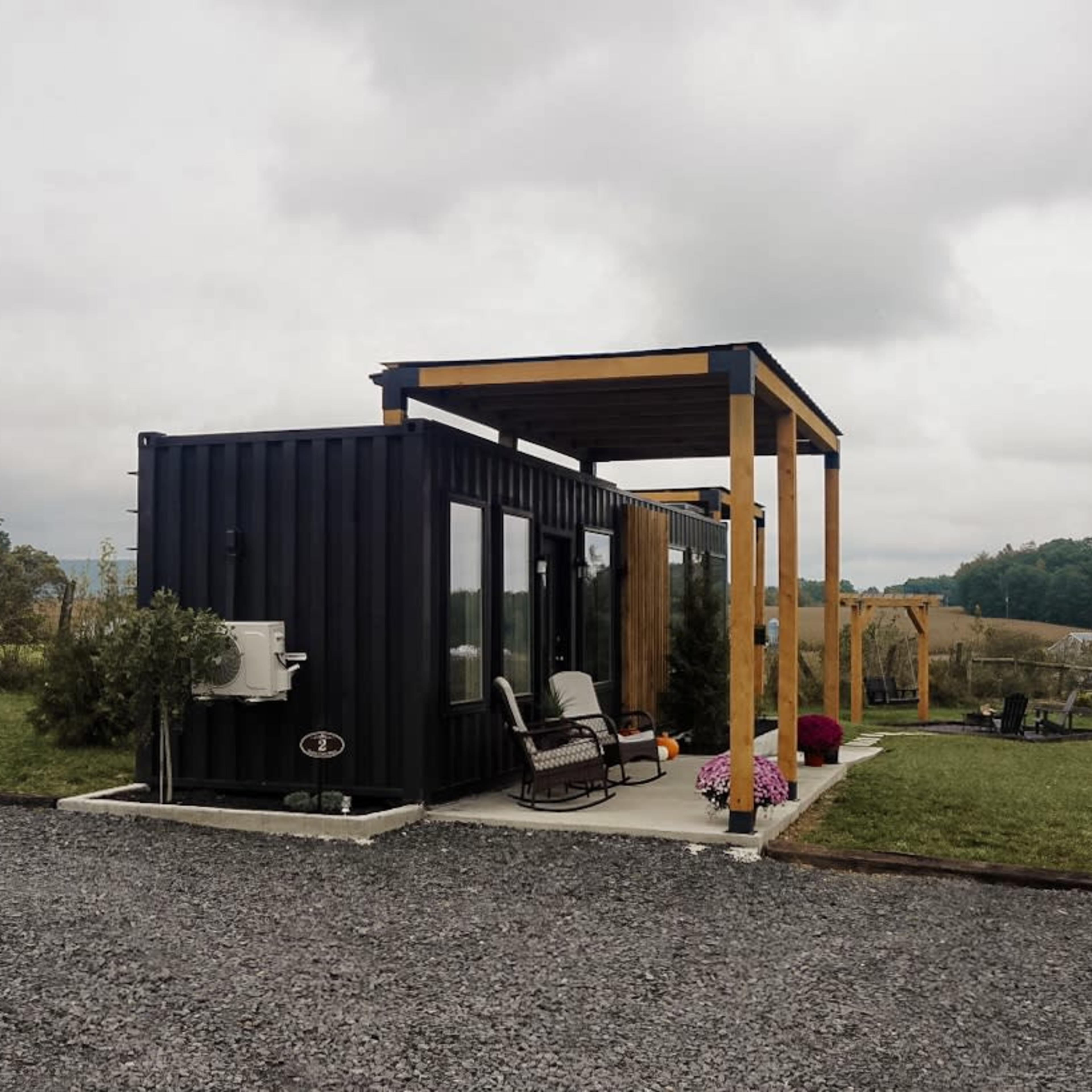 A modern black shipping container home is set on a gravel driveway with a covered porch, surrounded by a grassy landscape and distant hills.