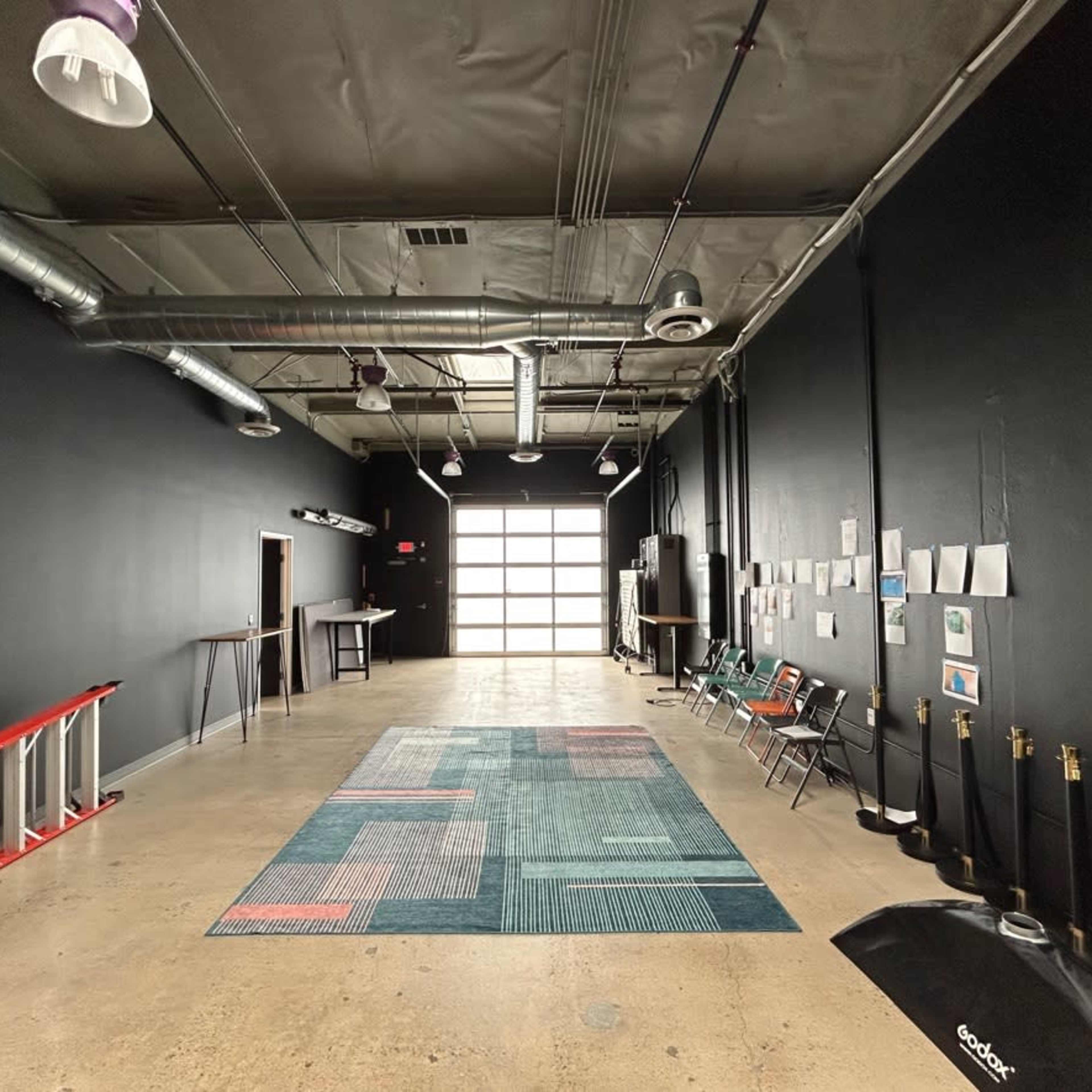 An empty industrial space with a concrete floor, black walls, a large garage door at one end, and various furnishings including chairs and a rug.