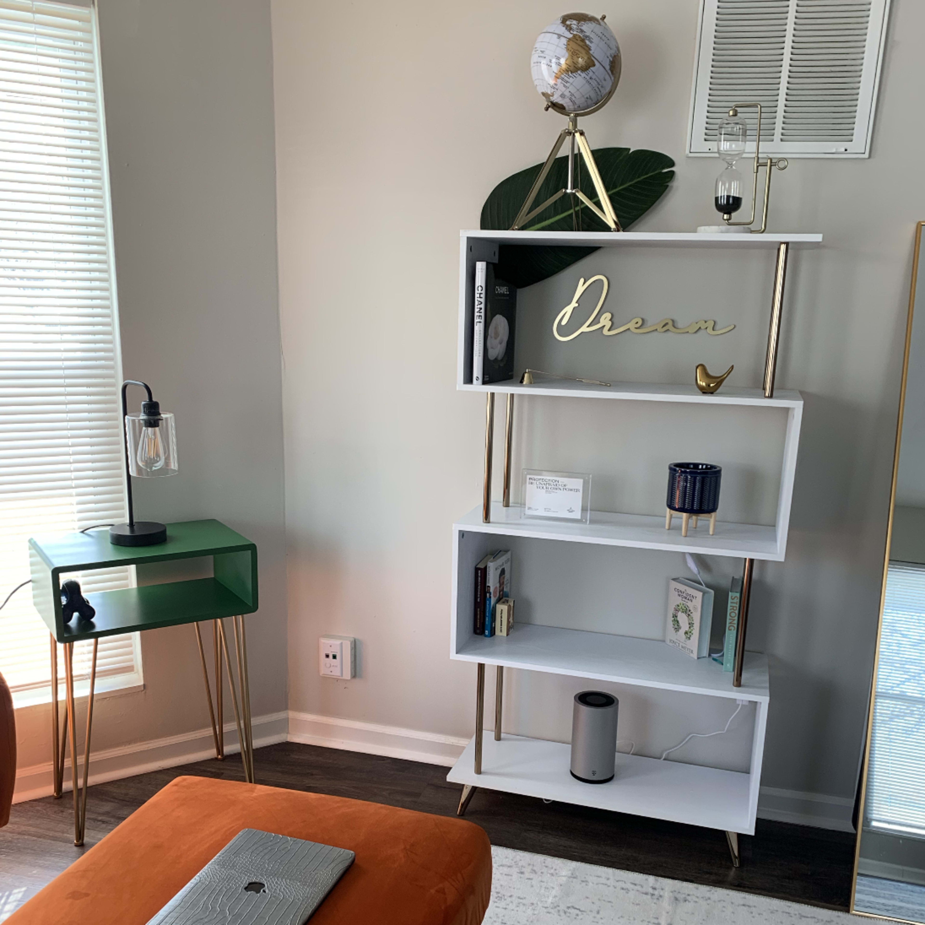 A modern living space featuring a white bookshelf with decor items, an orange ottoman, and a green side table with a lamp.