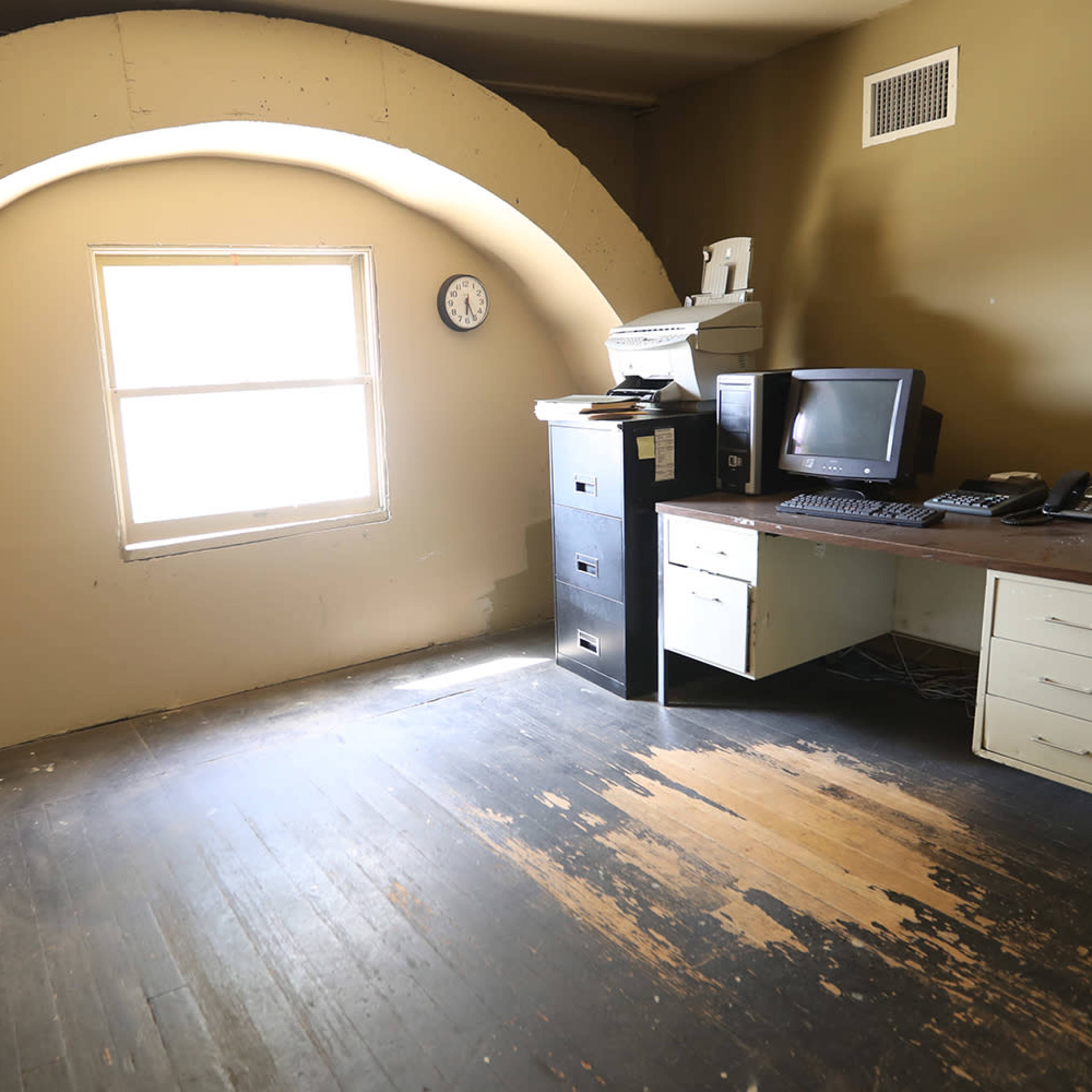 A sparsely furnished room with an arched window, a desk with a computer, a printer, and a filing cabinet.