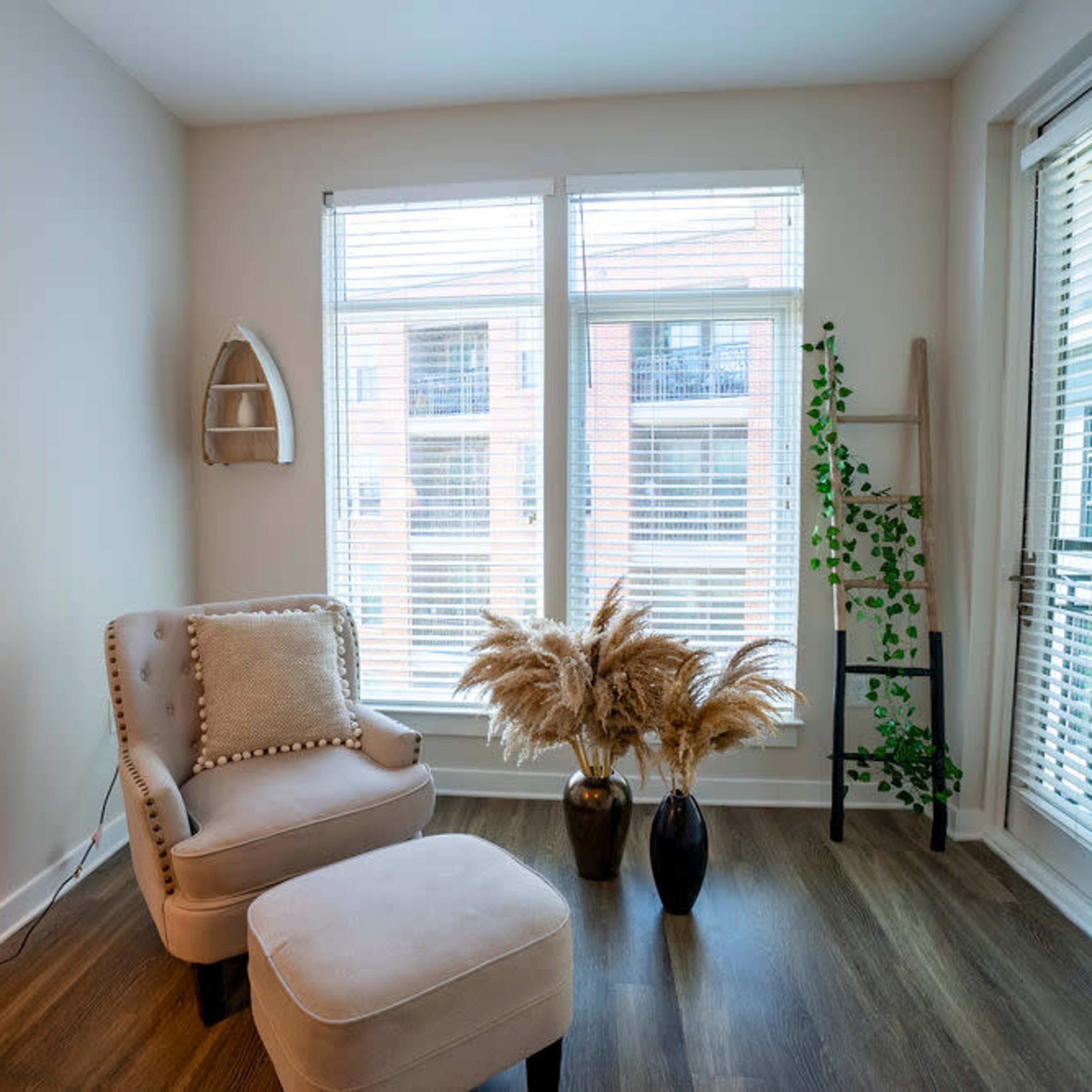 A cozy corner of a room featuring a chair and ottoman, two large windows with blinds, and decorative plants in a vase.