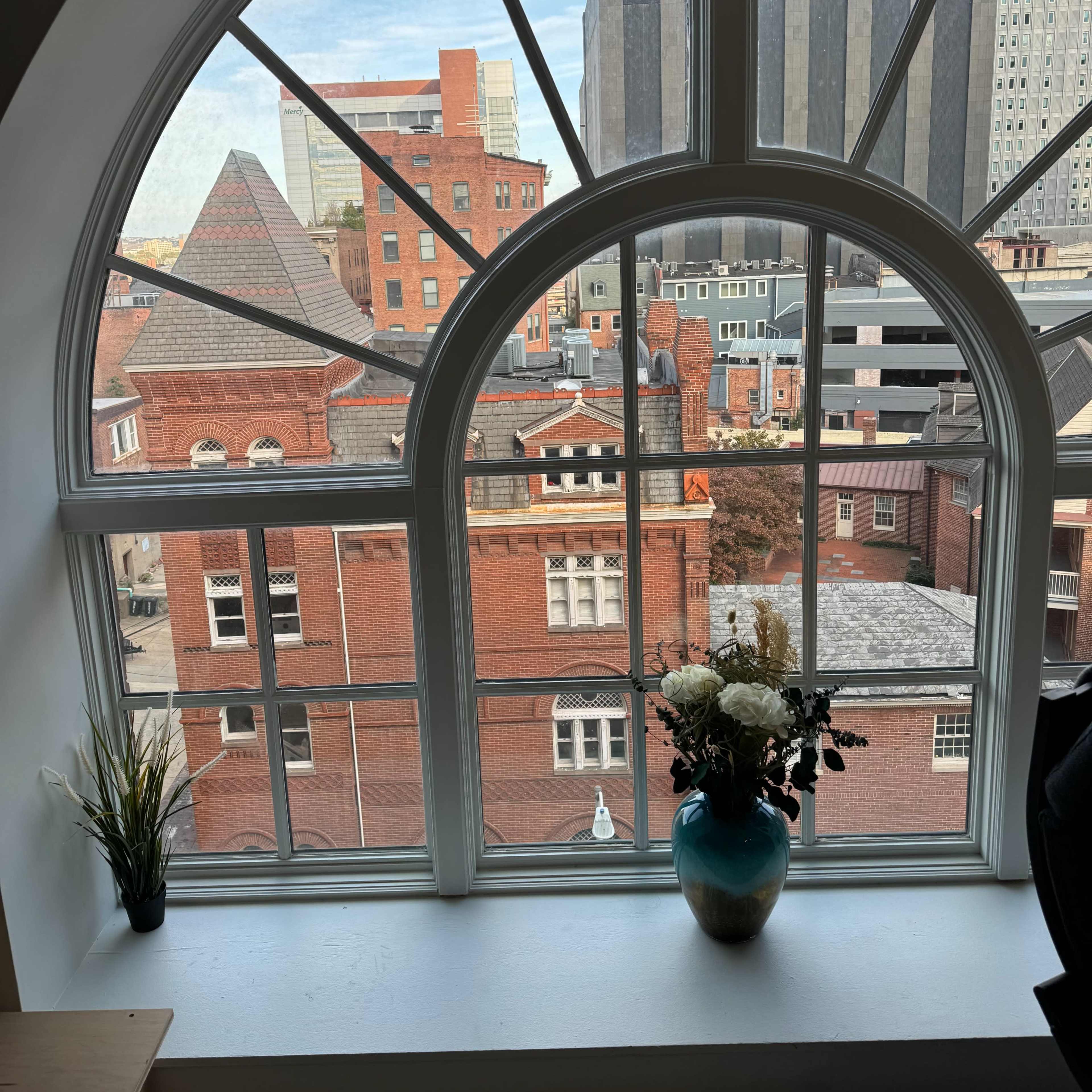 A large arched window framing a view of city buildings, with a small potted plant and a vase of flowers on the windowsill.