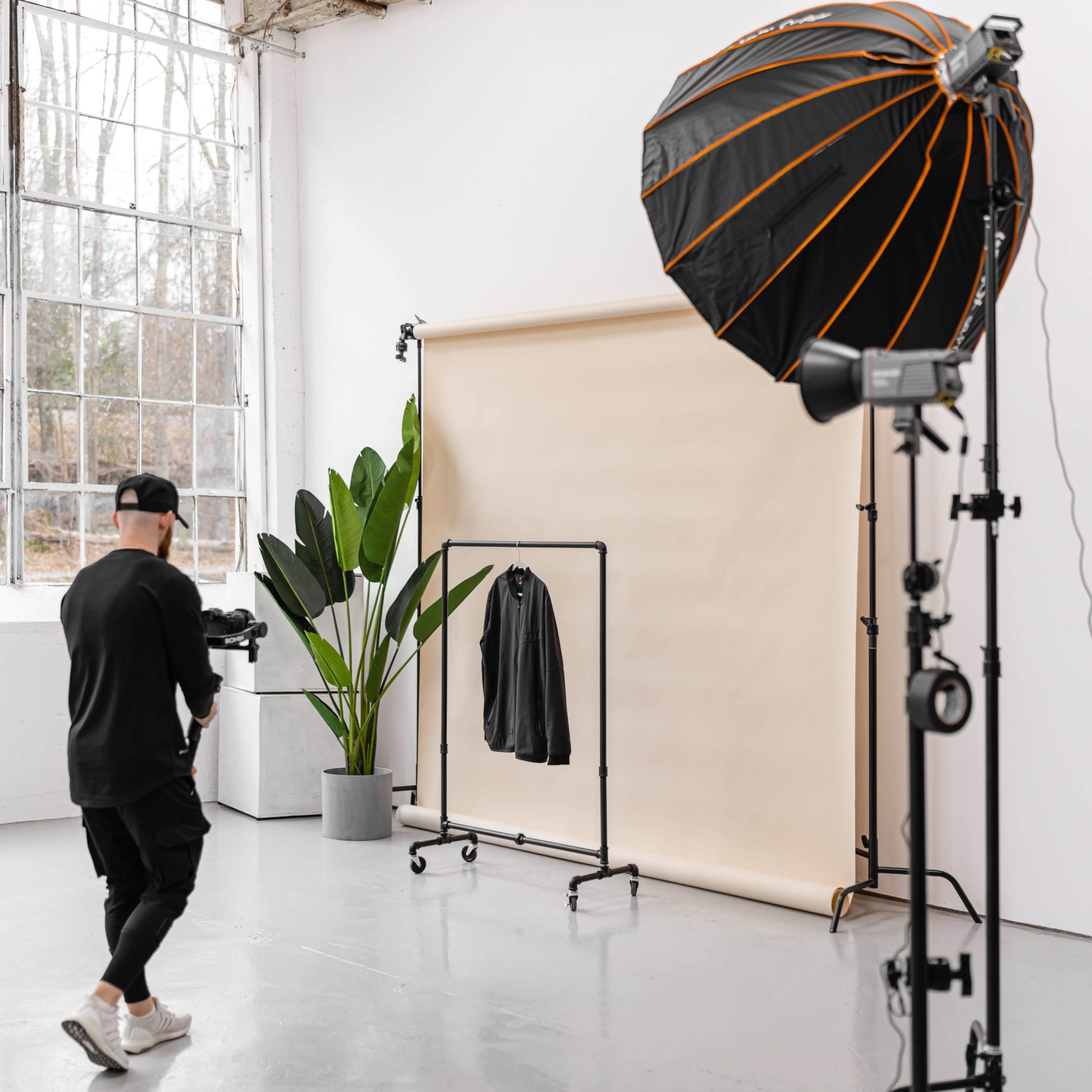 A photographer stands in a studio with a backdrop and a clothing rack holding a jacket, accompanied by a large softbox light and a potted plant.