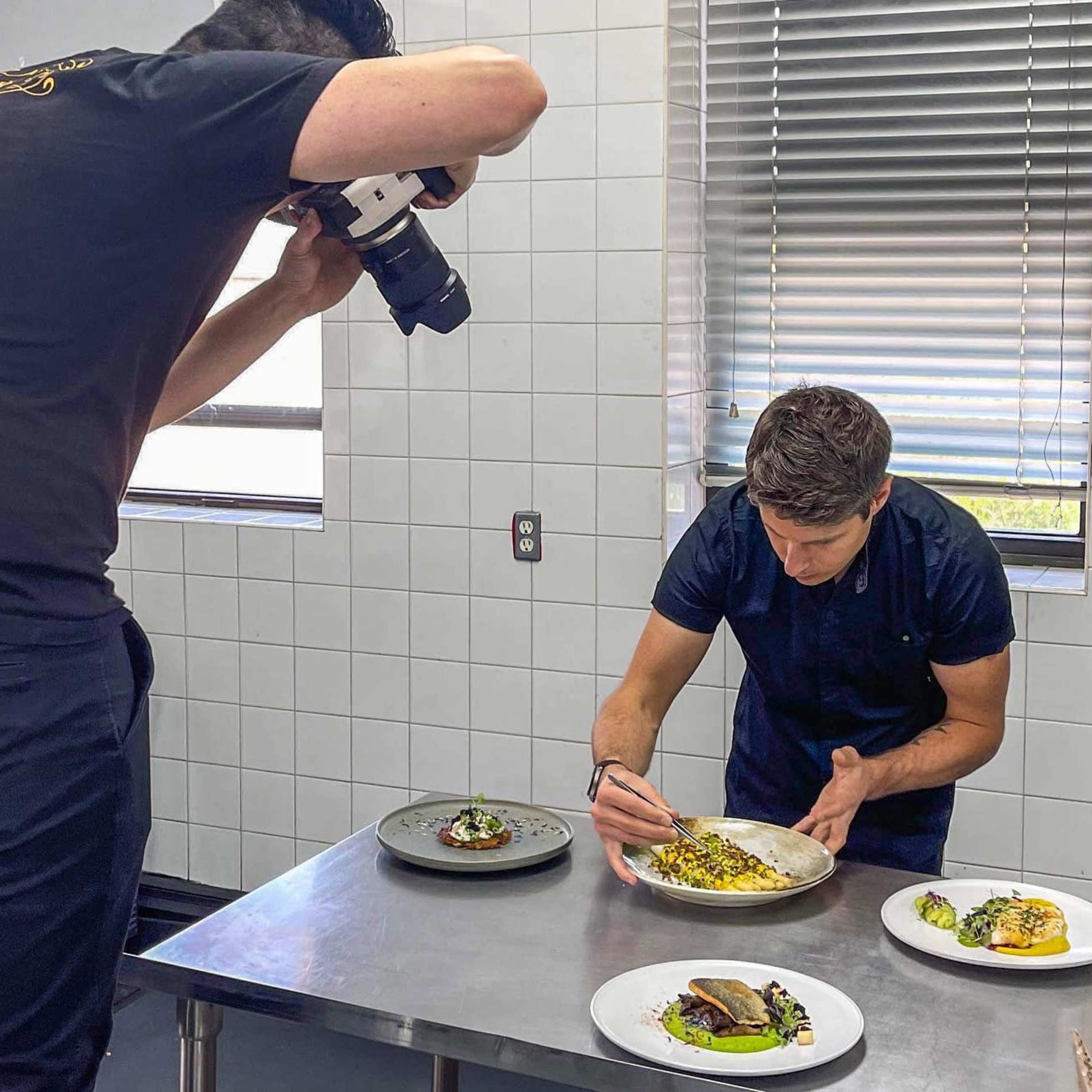 A chef decorates a plate of food while a photographer captures the scene in a brightly lit kitchen.