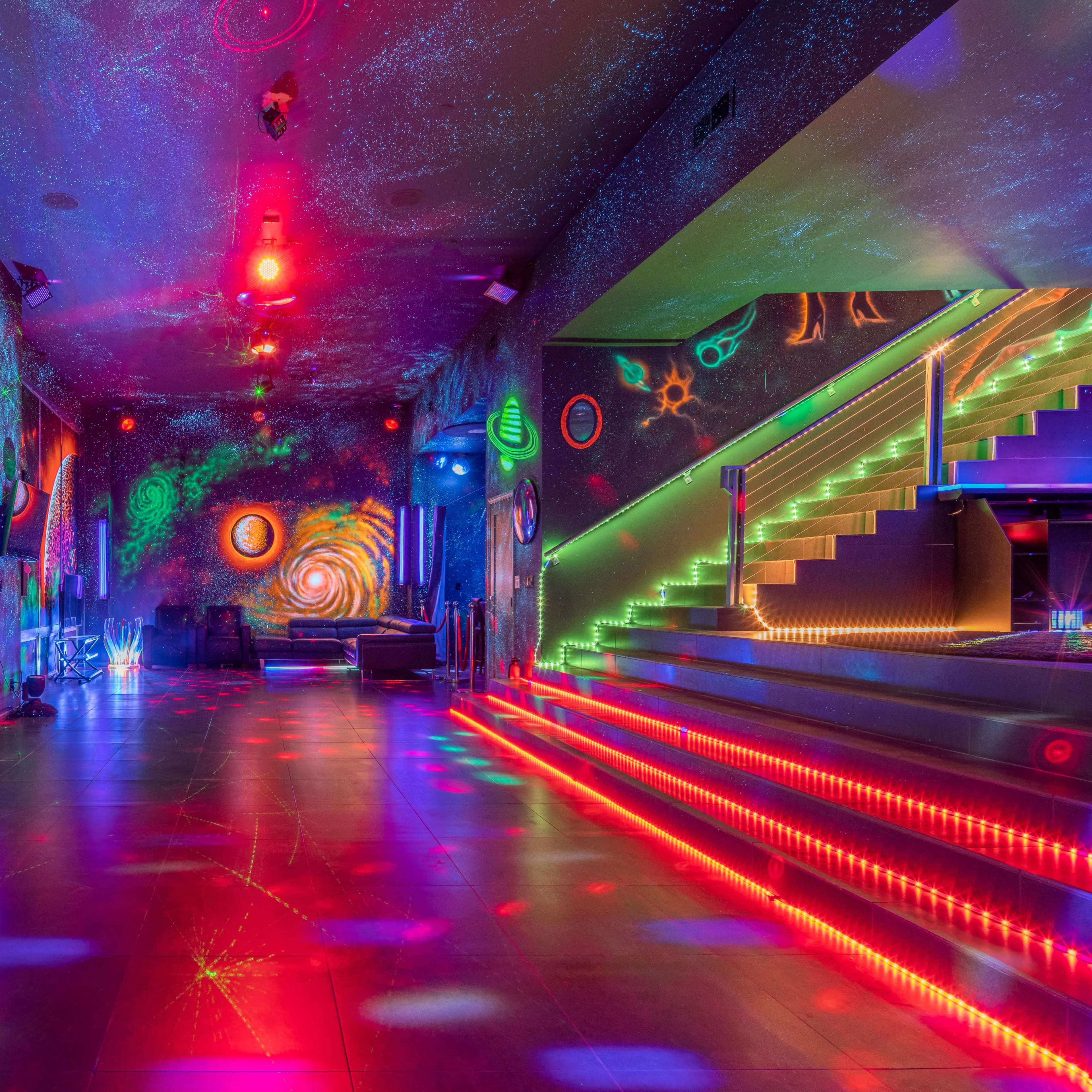 The interior of a brightly lit lounge features colorful, psychedelic wall art and illuminated stairs with vibrant light patterns on the floor.
