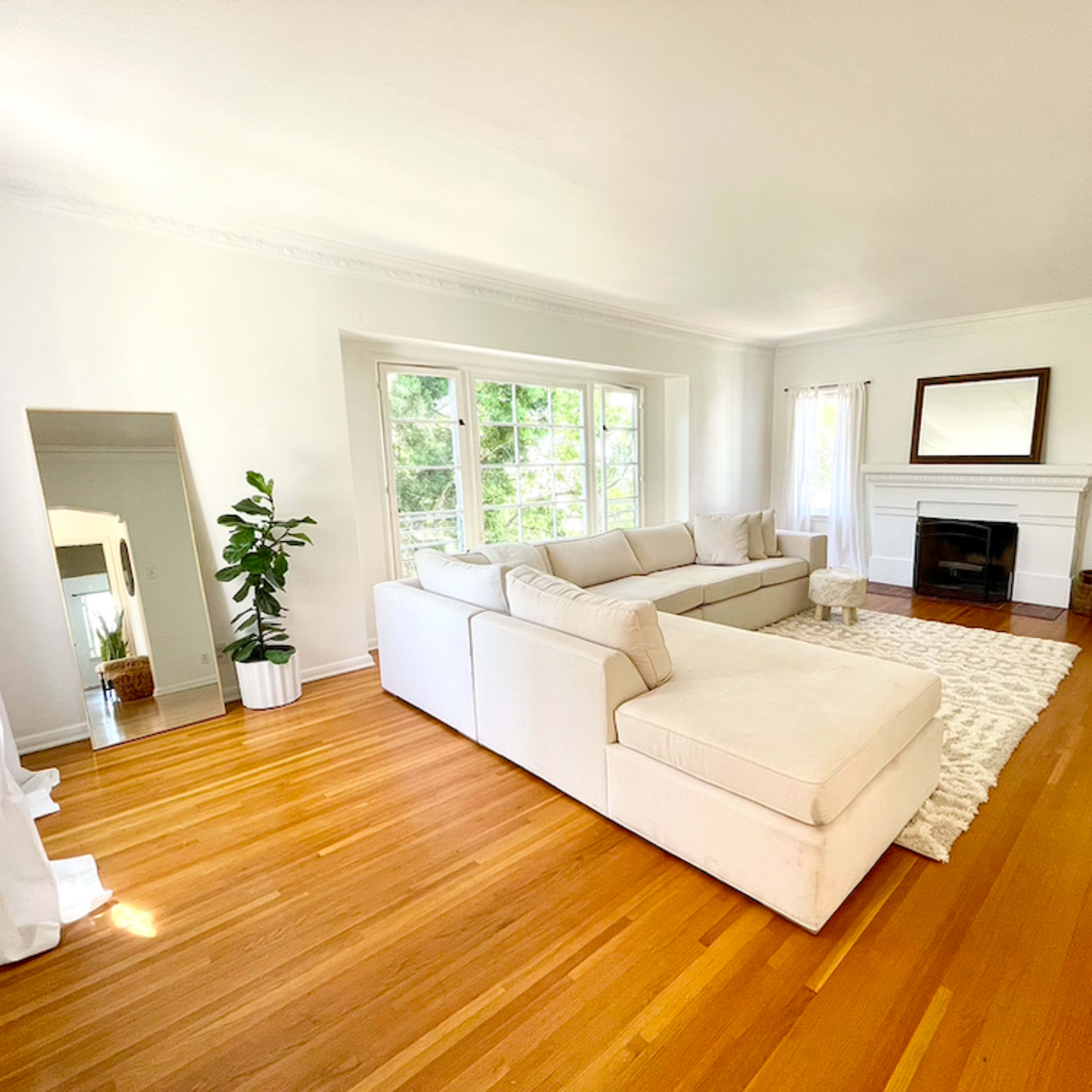 A spacious living room features a light-colored sectional sofa, hardwood floors, a fireplace, and large windows with natural light.
