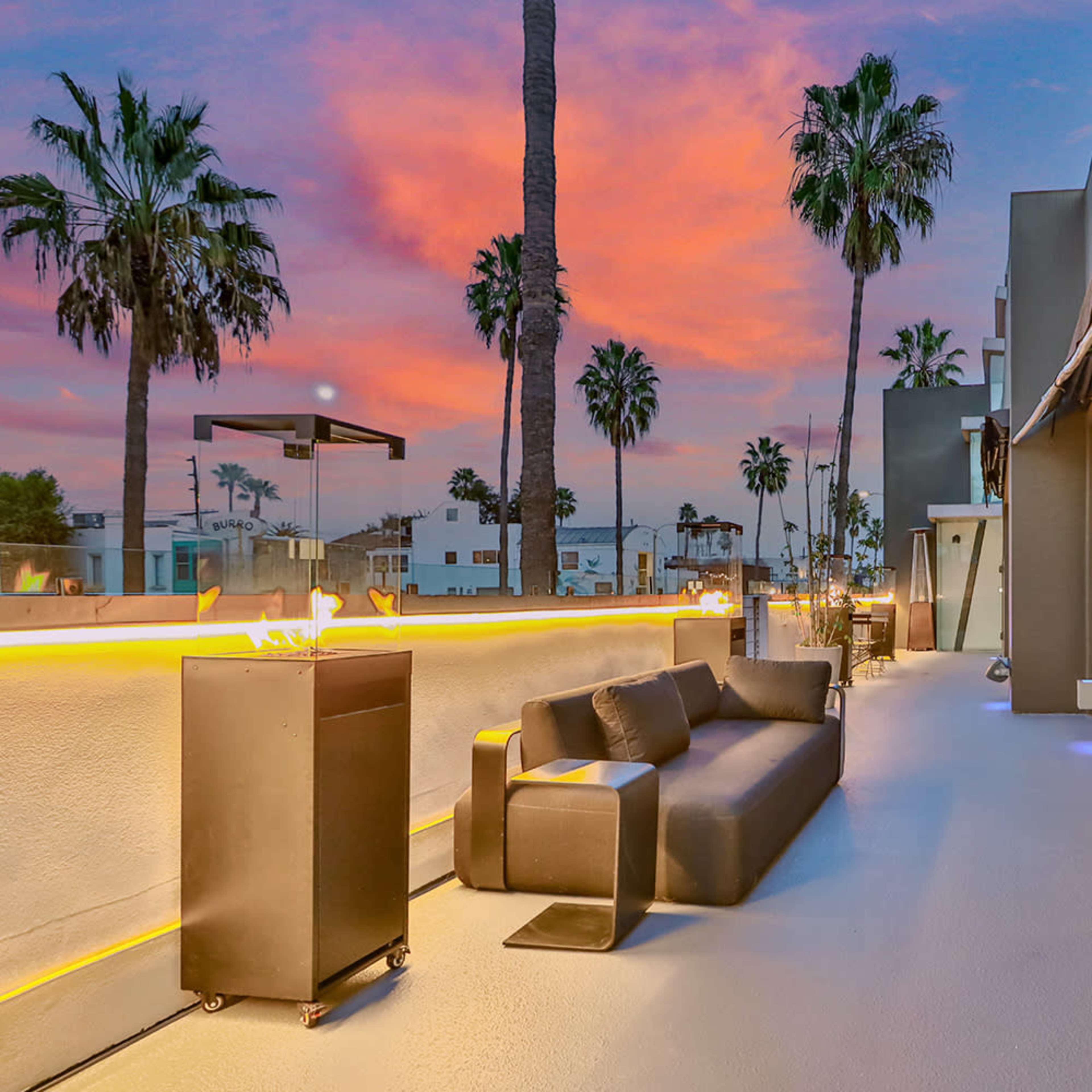 A balcony area with a dark sofa and palm trees silhouetted against a colorful sunset sky.
