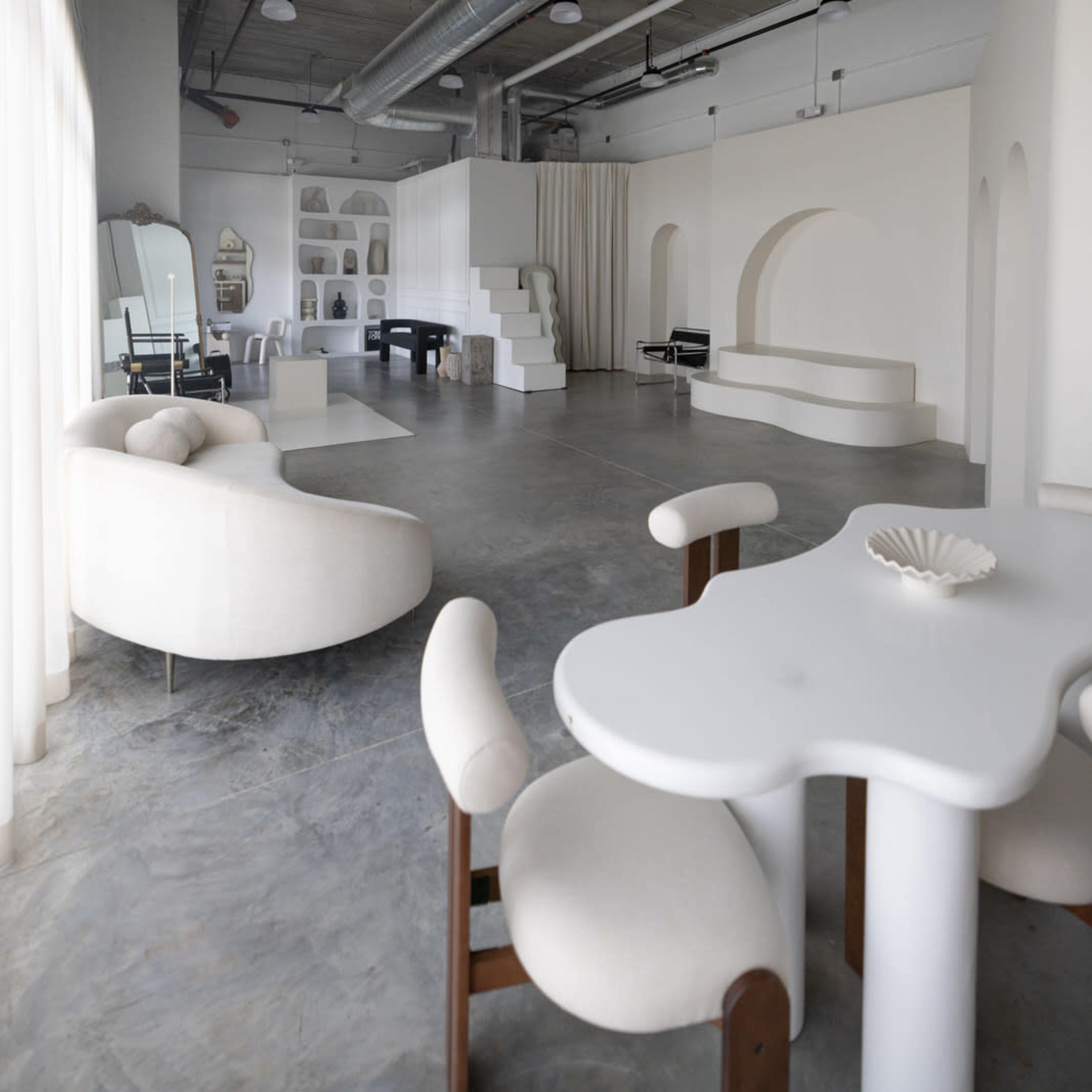 A modern interior space with minimalist furniture, including a curved sofa and a unique table, set against a backdrop of white walls and concrete flooring.
