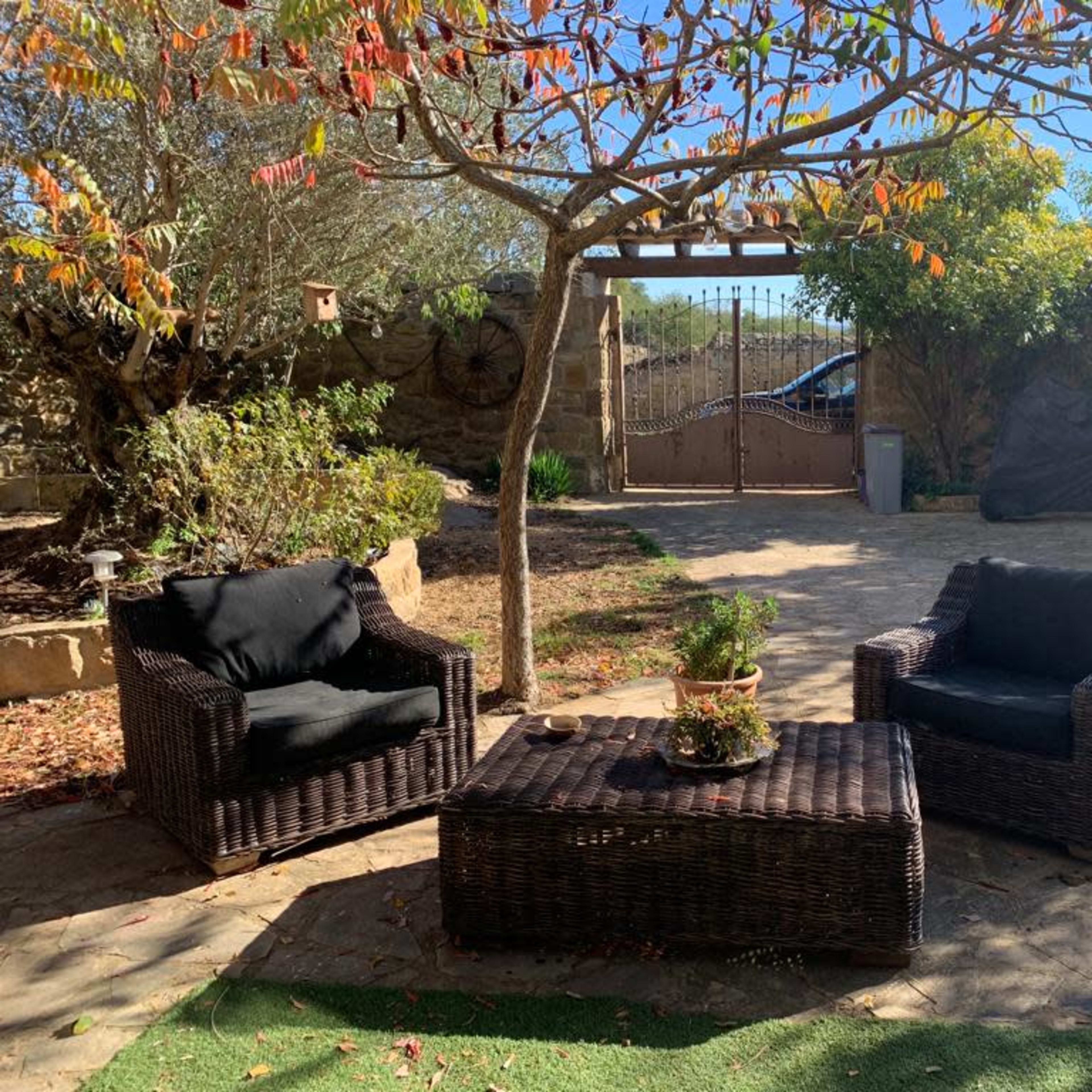 A patio area features two wicker chairs and a table surrounded by trees and a gate in the background.