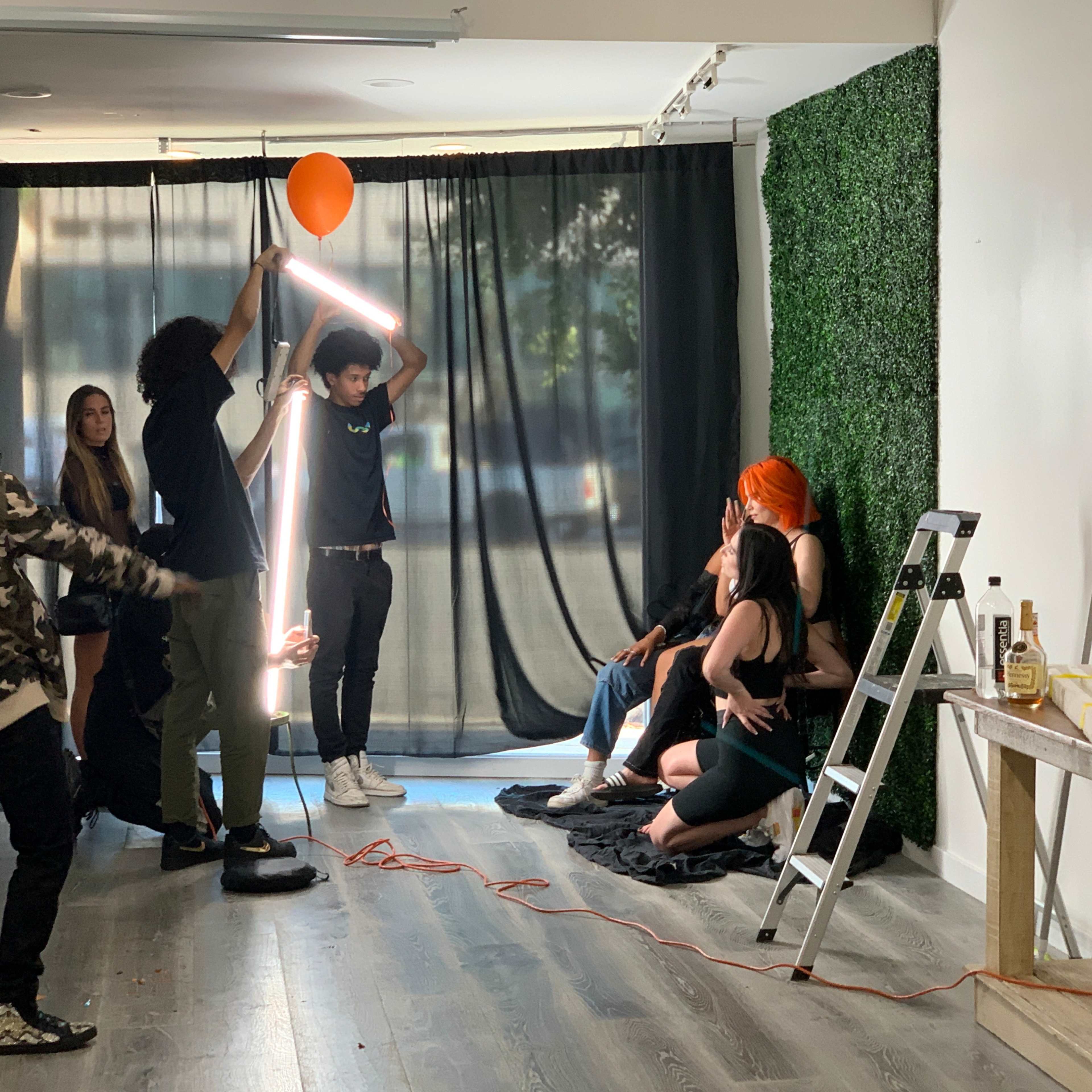 A group of people poses for a photo shoot in a studio, with a person holding a neon light and another holding a balloon.