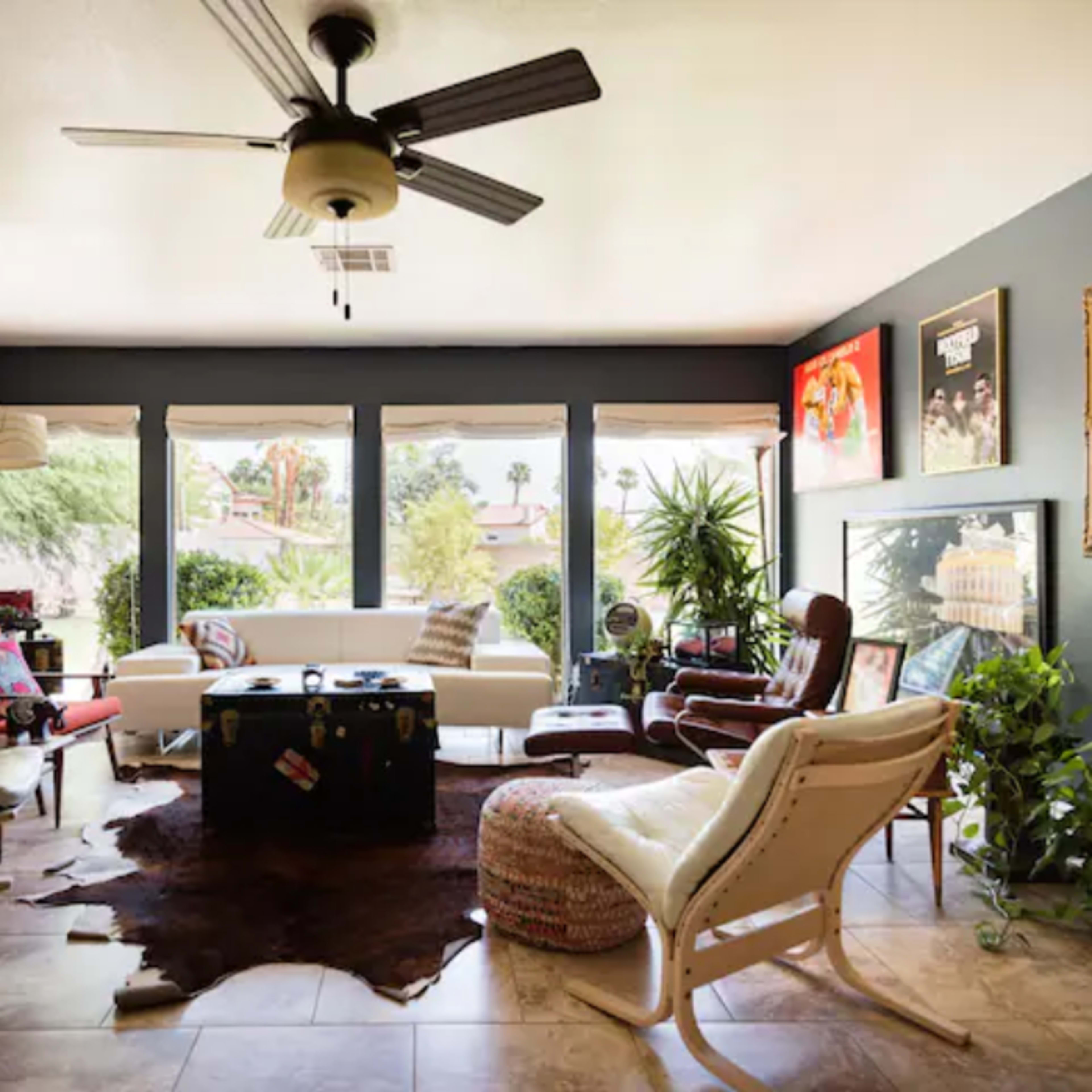 A bright living room with various seating options, a coffee table, and large windows that offer a view of greenery outside.