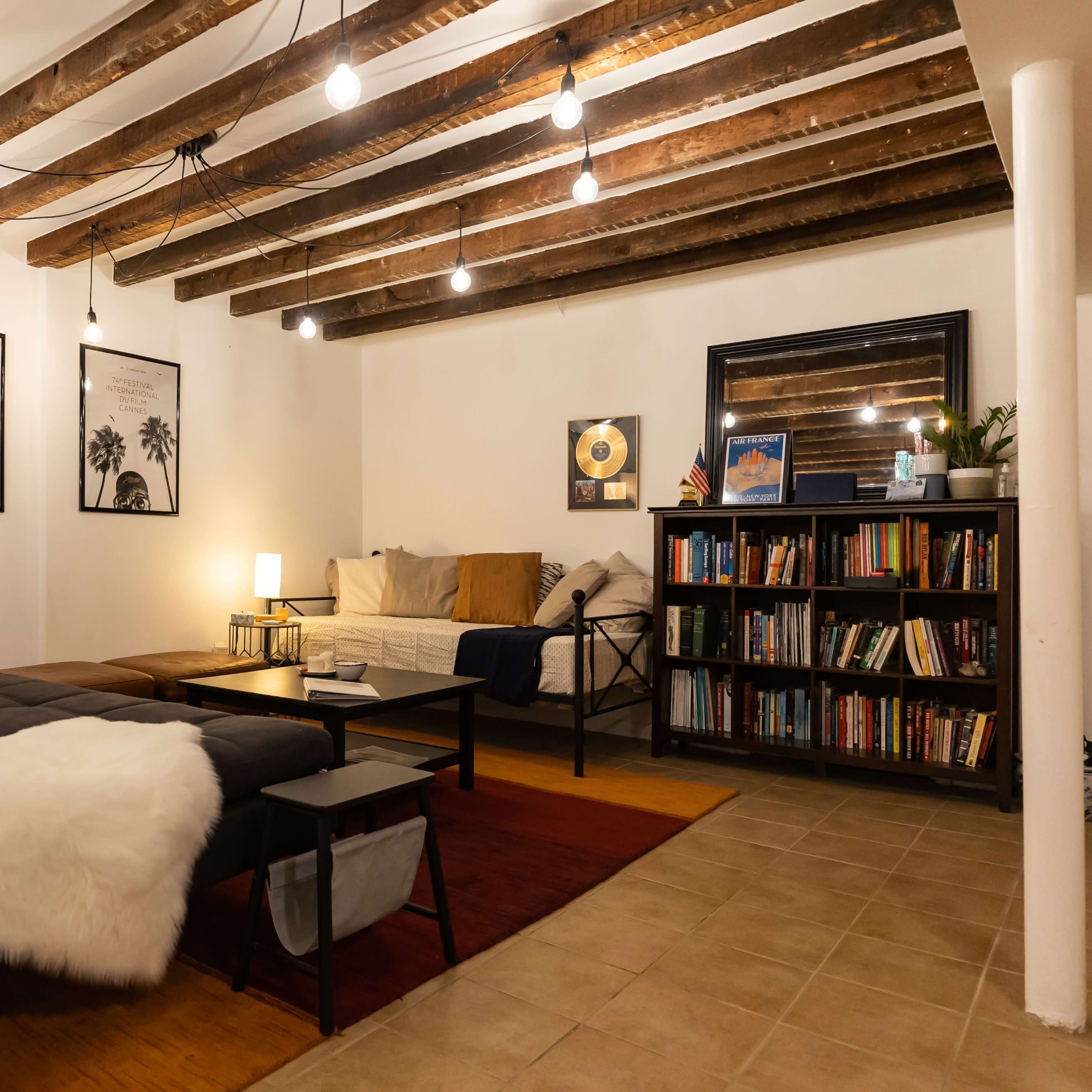 A cozy room featuring a gray chaise lounge, a wooden bookshelf filled with books, and a seating area with a coffee table, illuminated by hanging light bulbs and a floor lamp.