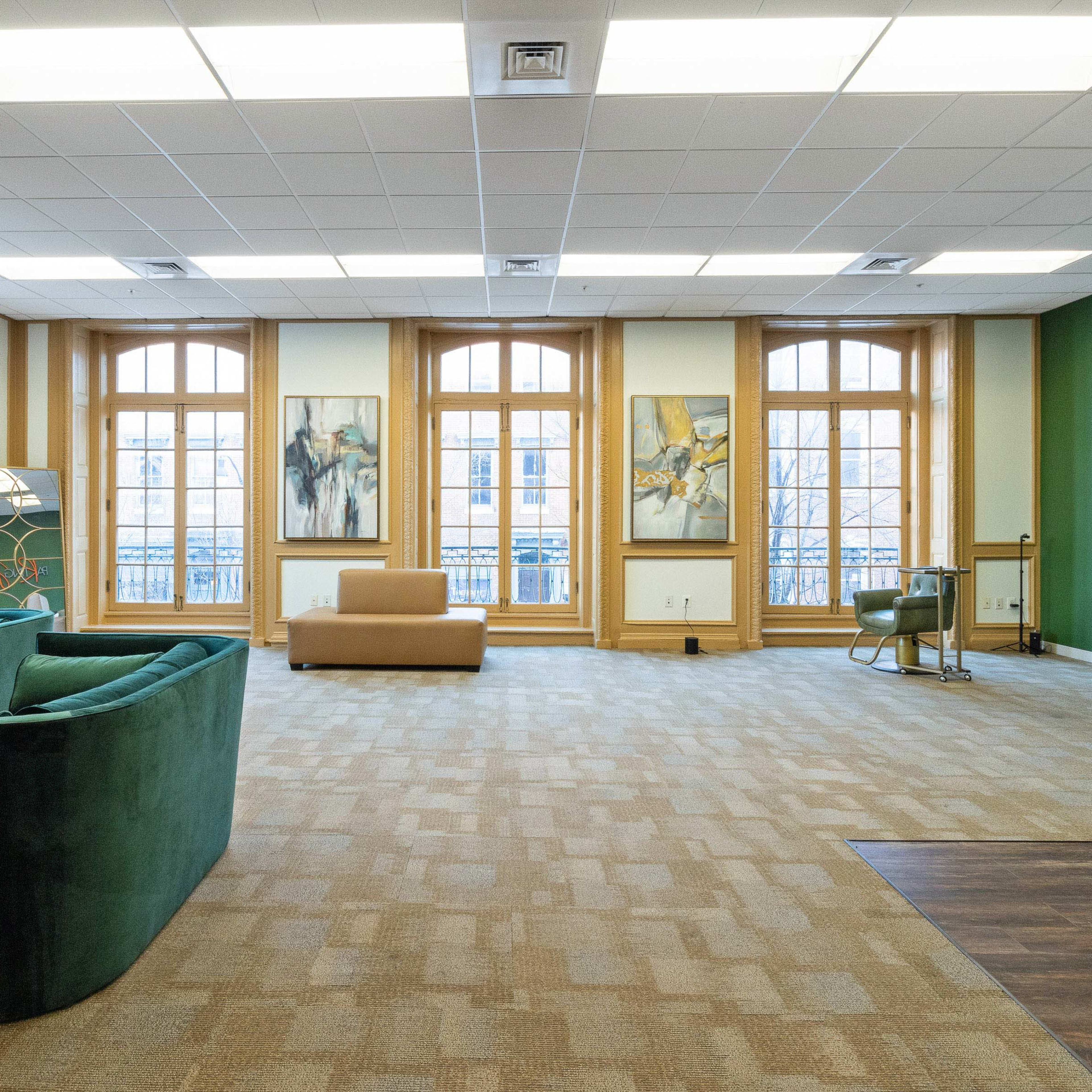 A spacious room featuring large windows, a green sofa, a beige couch, and abstract artwork on the walls.