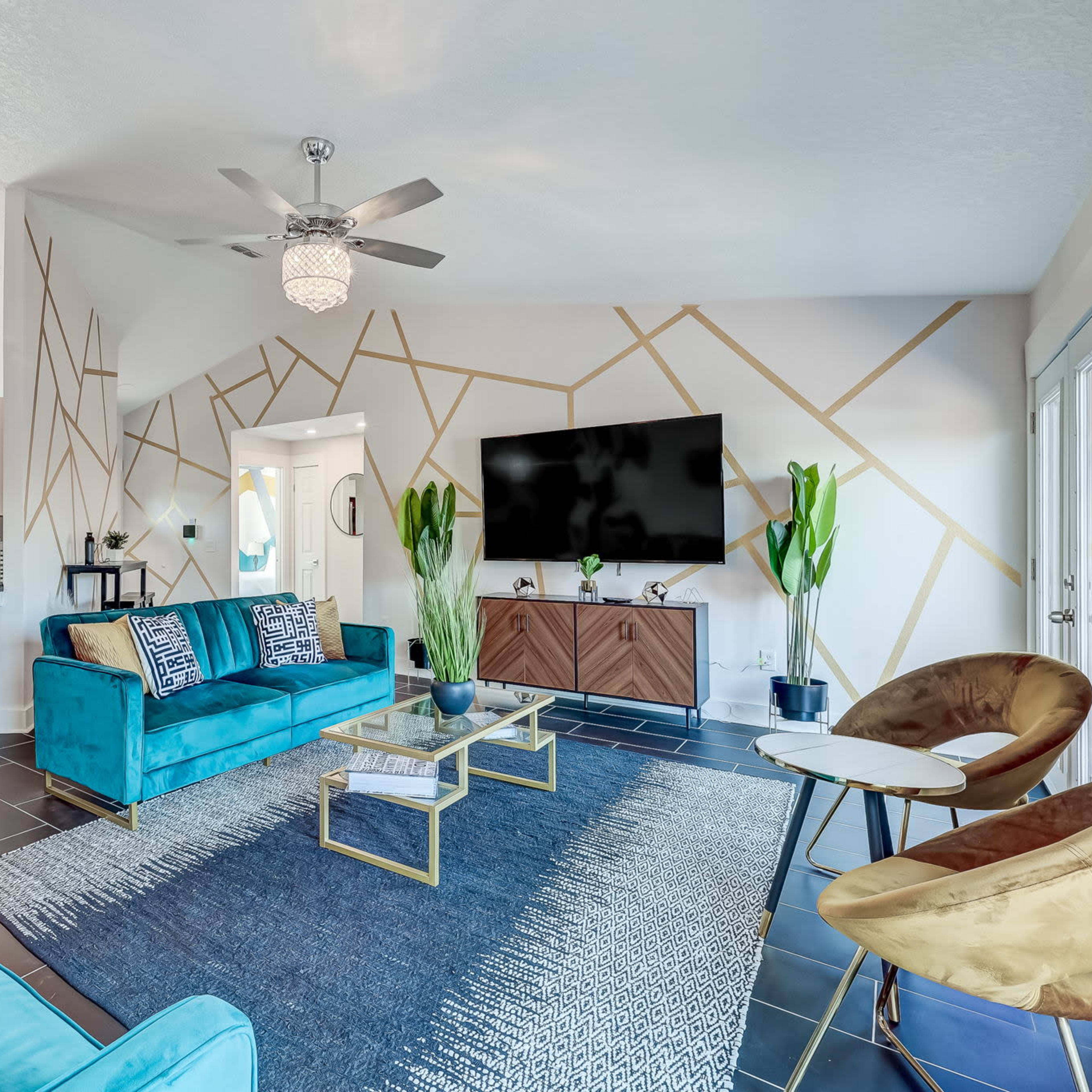 The living room features turquoise sofas, a geometric accent wall, a large television, and decorative plants.