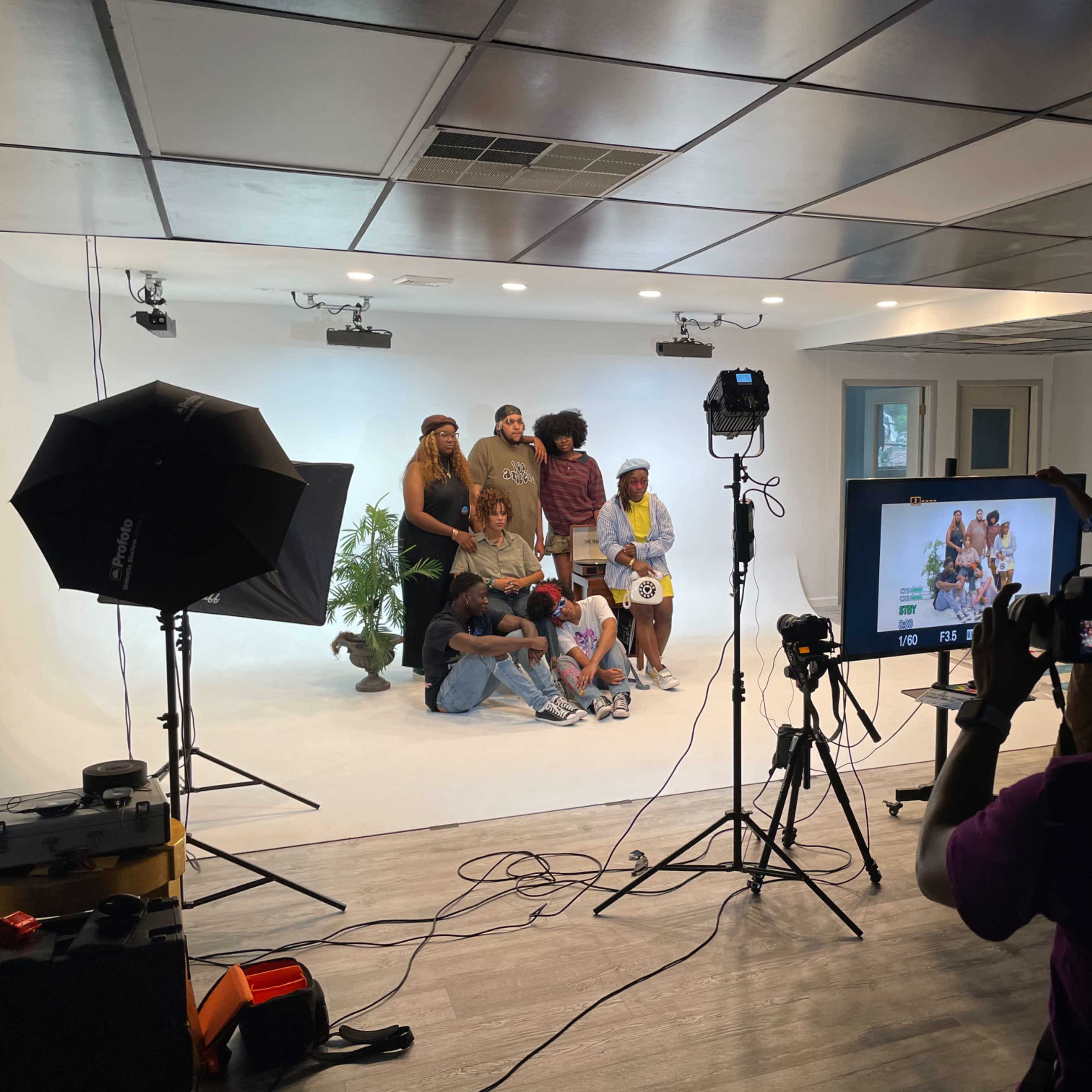 A group of people poses for a photo in a studio with a white backdrop, while others set up cameras and lighting equipment.