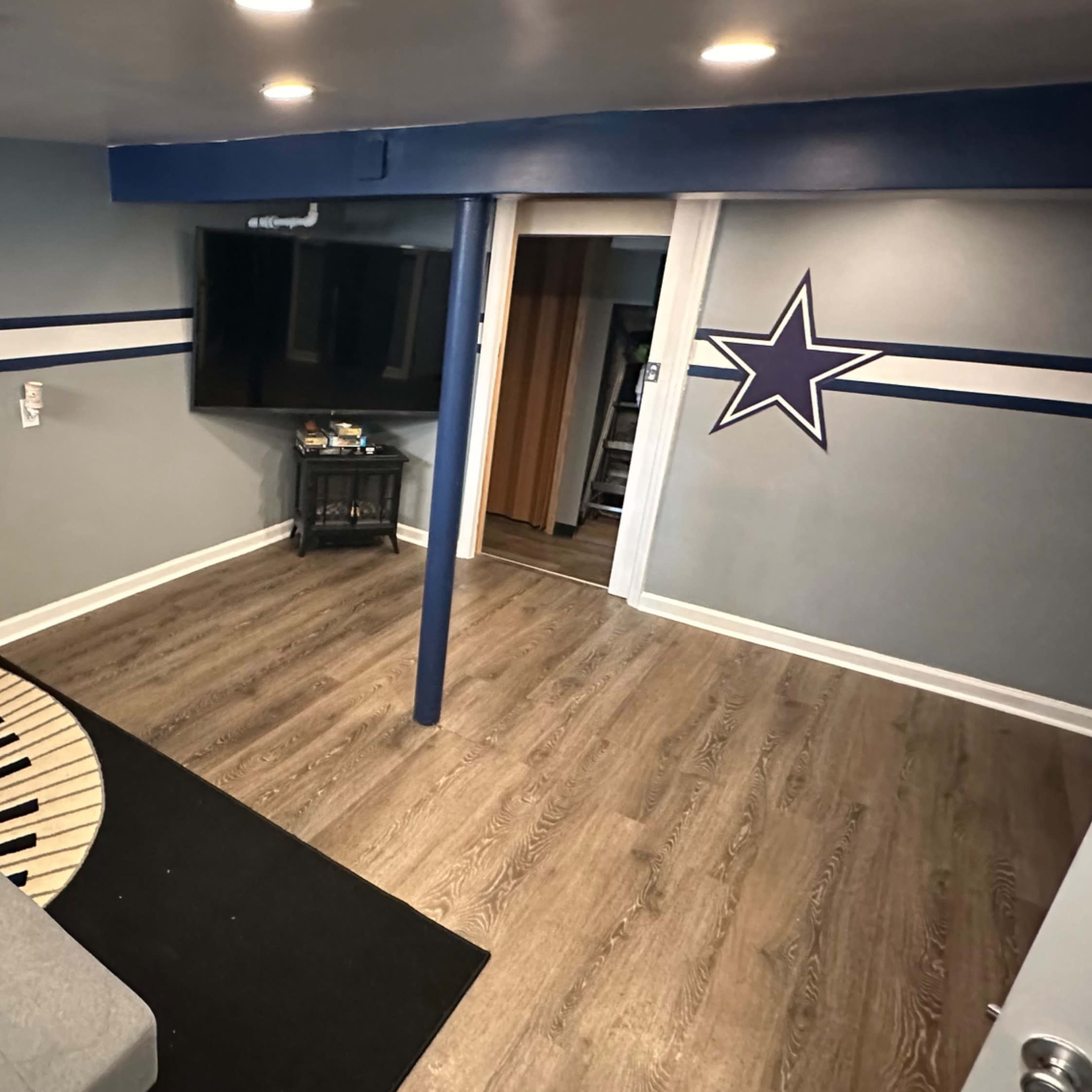 A basement room with a gray sofa, a black rug shaped like piano keys, a television mounted on the wall, and a star design on the wall.