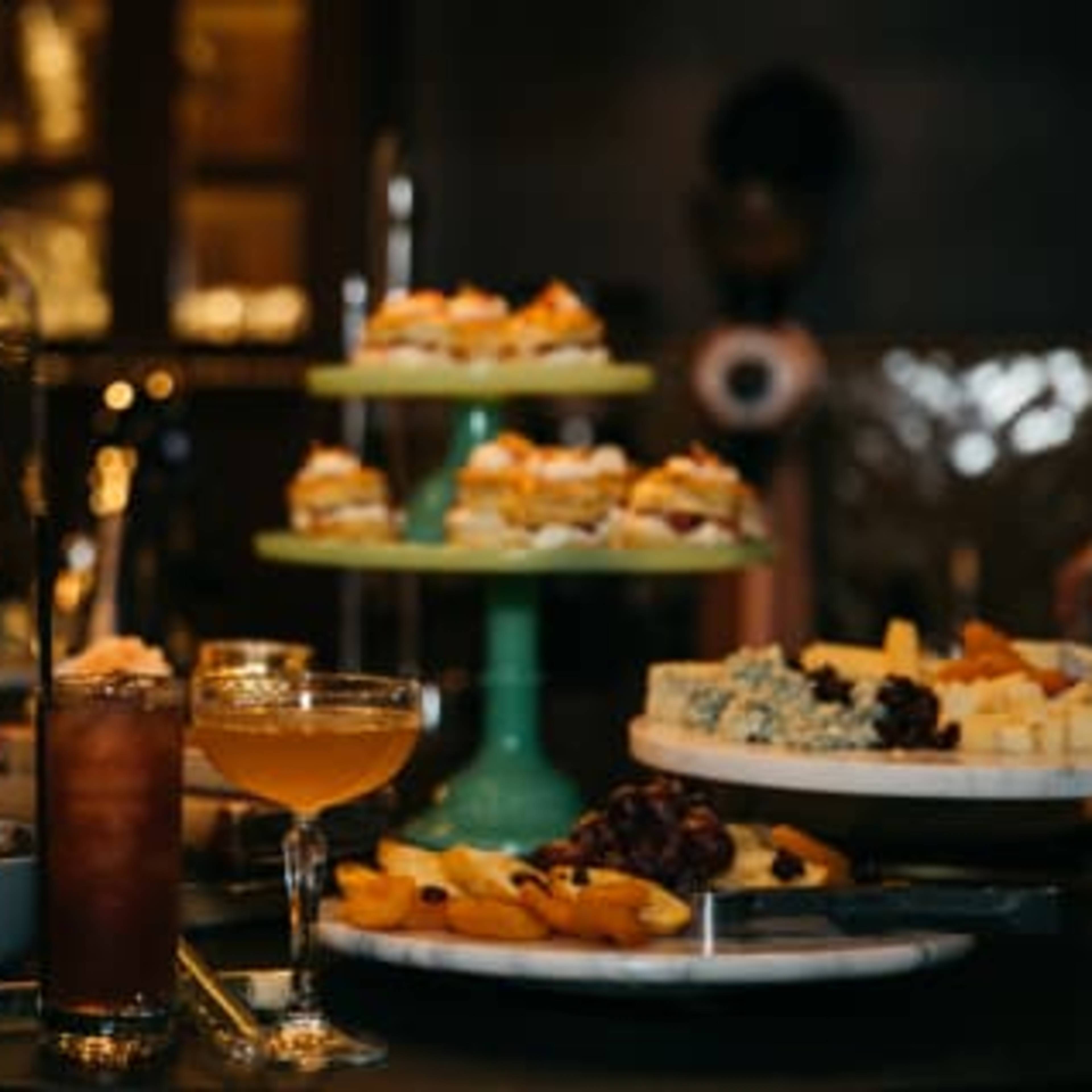 A buffet table displays an array of appetizers, desserts on stands, and drinks in glasses.