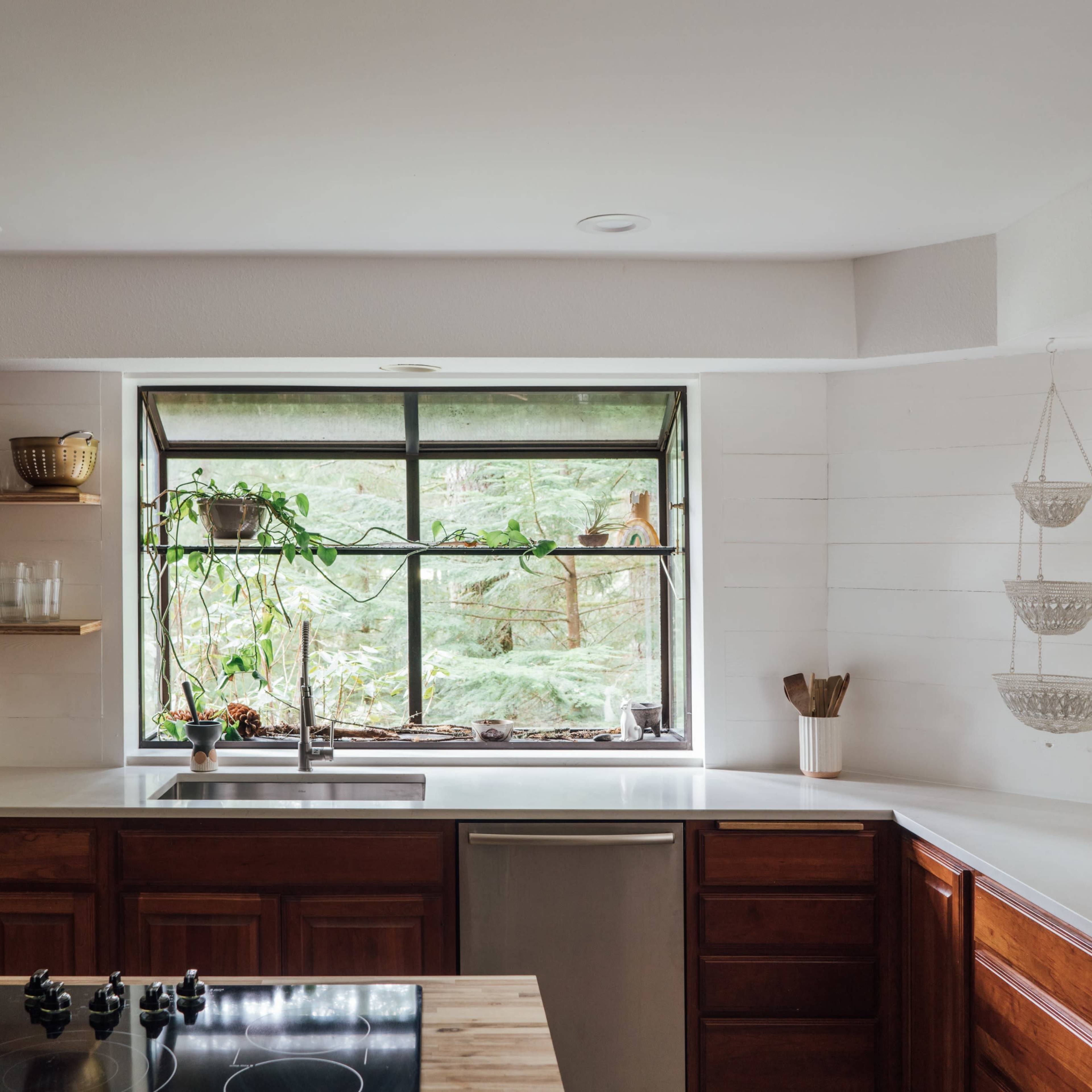 A modern kitchen features wooden cabinets, a stainless steel sink beneath a large window, and a countertop with a built-in stove.