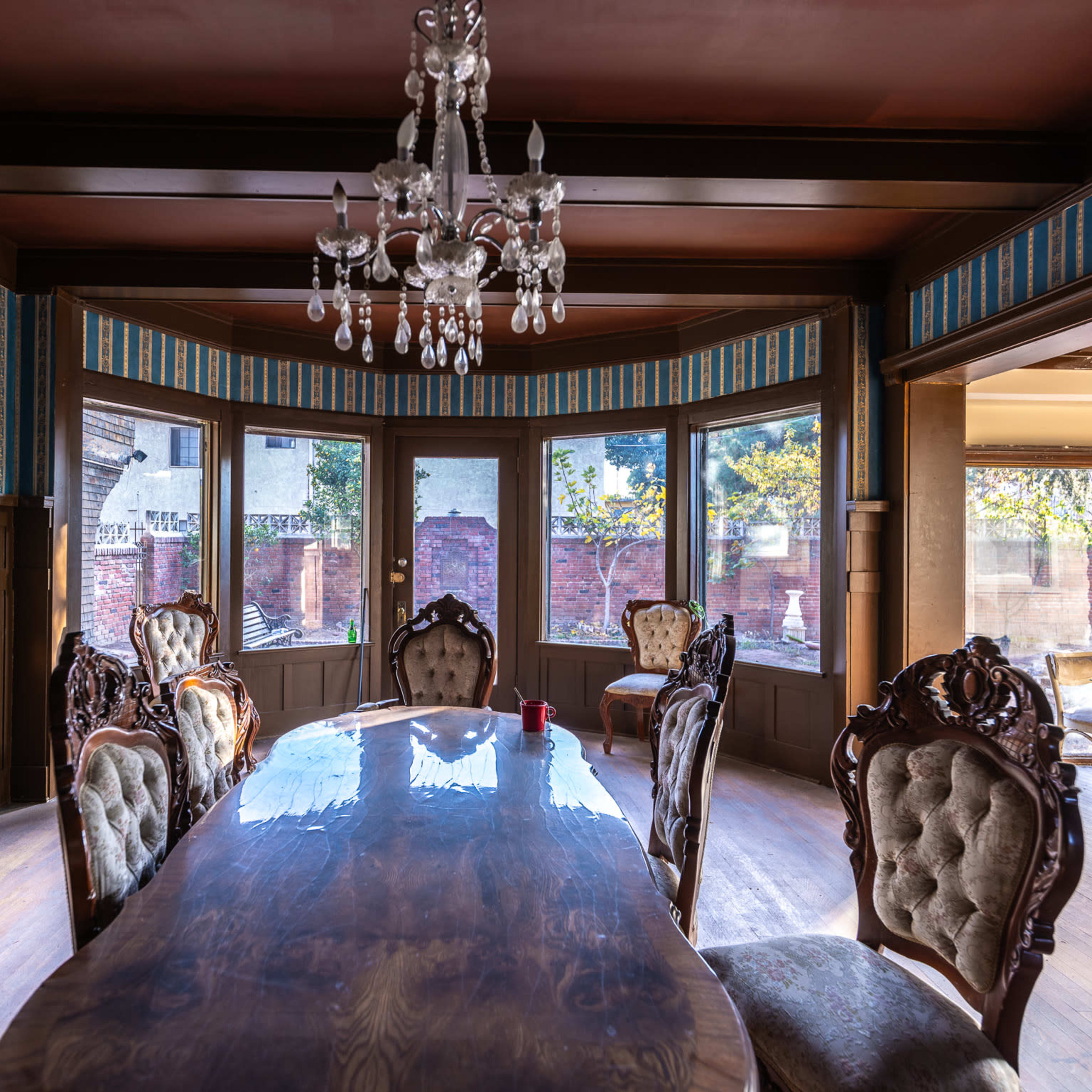 A dining room with a large wooden table and six upholstered chairs under a crystal chandelier, featuring patterned wallpaper and large windows overlooking a garden.