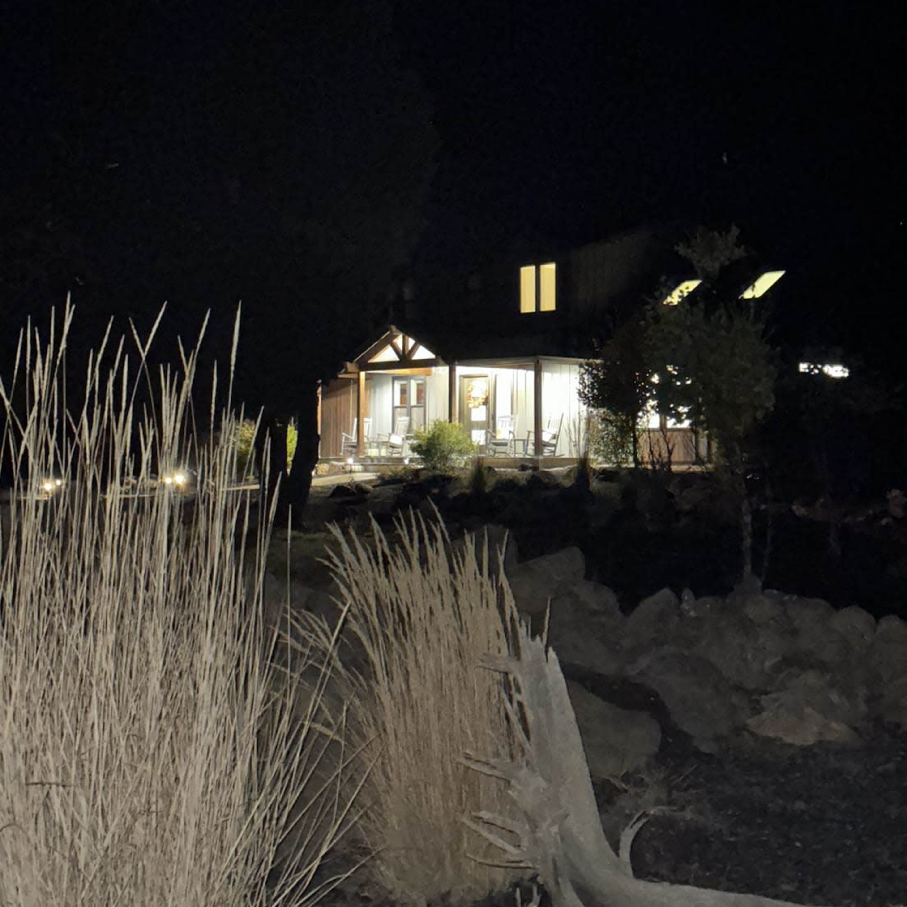 A well-lit house stands in a dark landscape, surrounded by rocks and dry grasses.