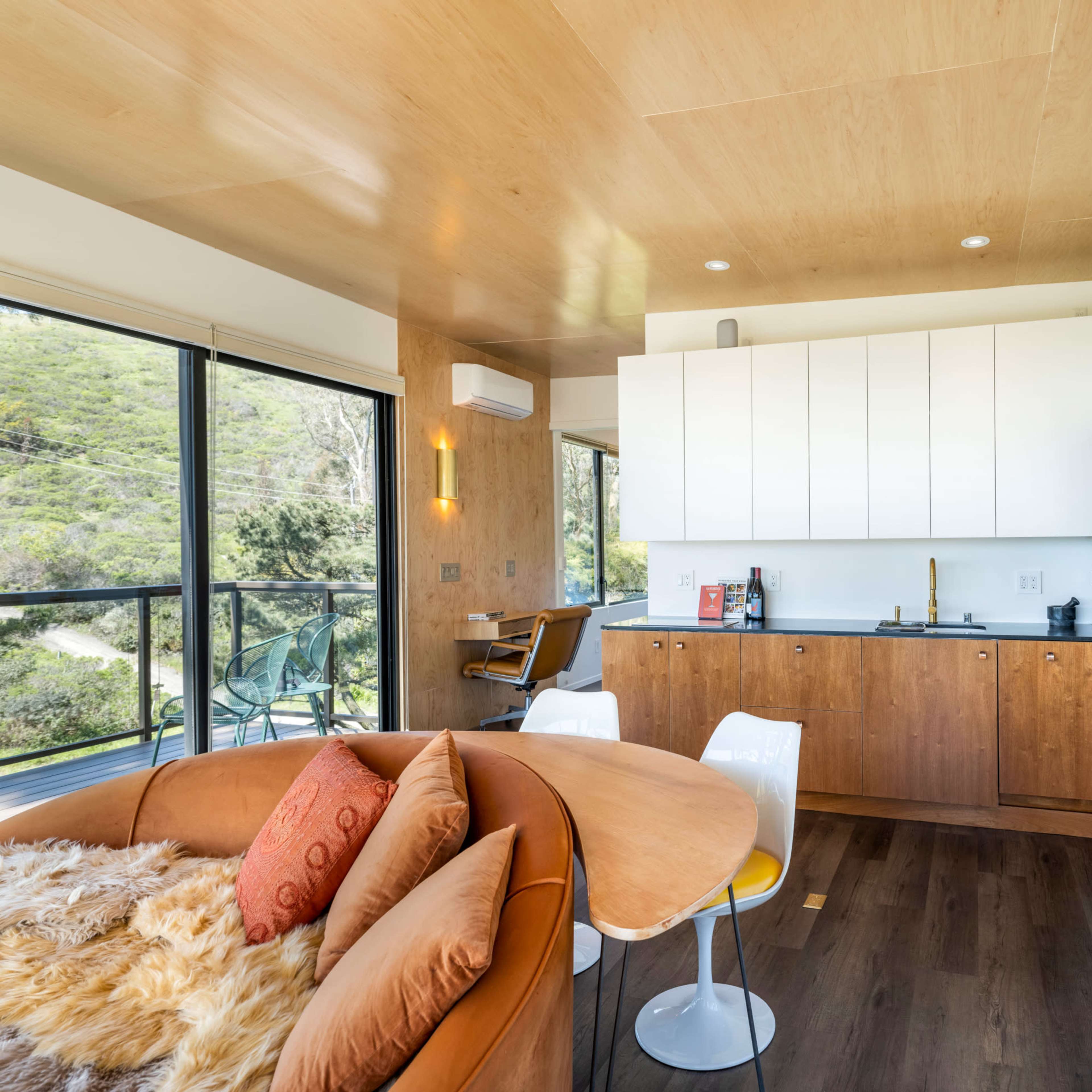 A modern, open-concept living space featuring a kitchen with white cabinets, a wooden dining table, and large windows overlooking greenery.