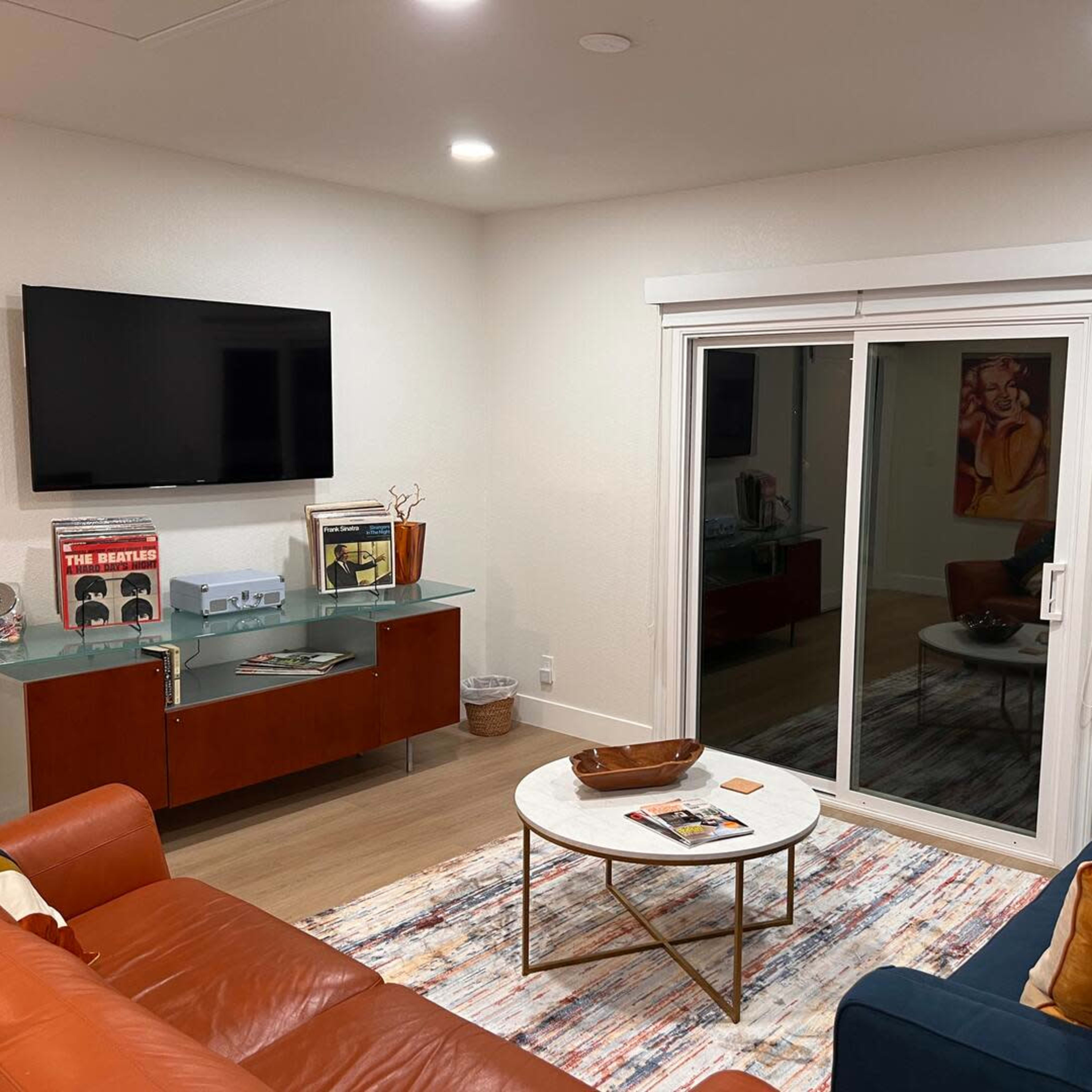 The room features a modern living area with a television mounted on the wall, a glass coffee table, and a sliding door leading outside, surrounded by a stylish couch and various decor items.