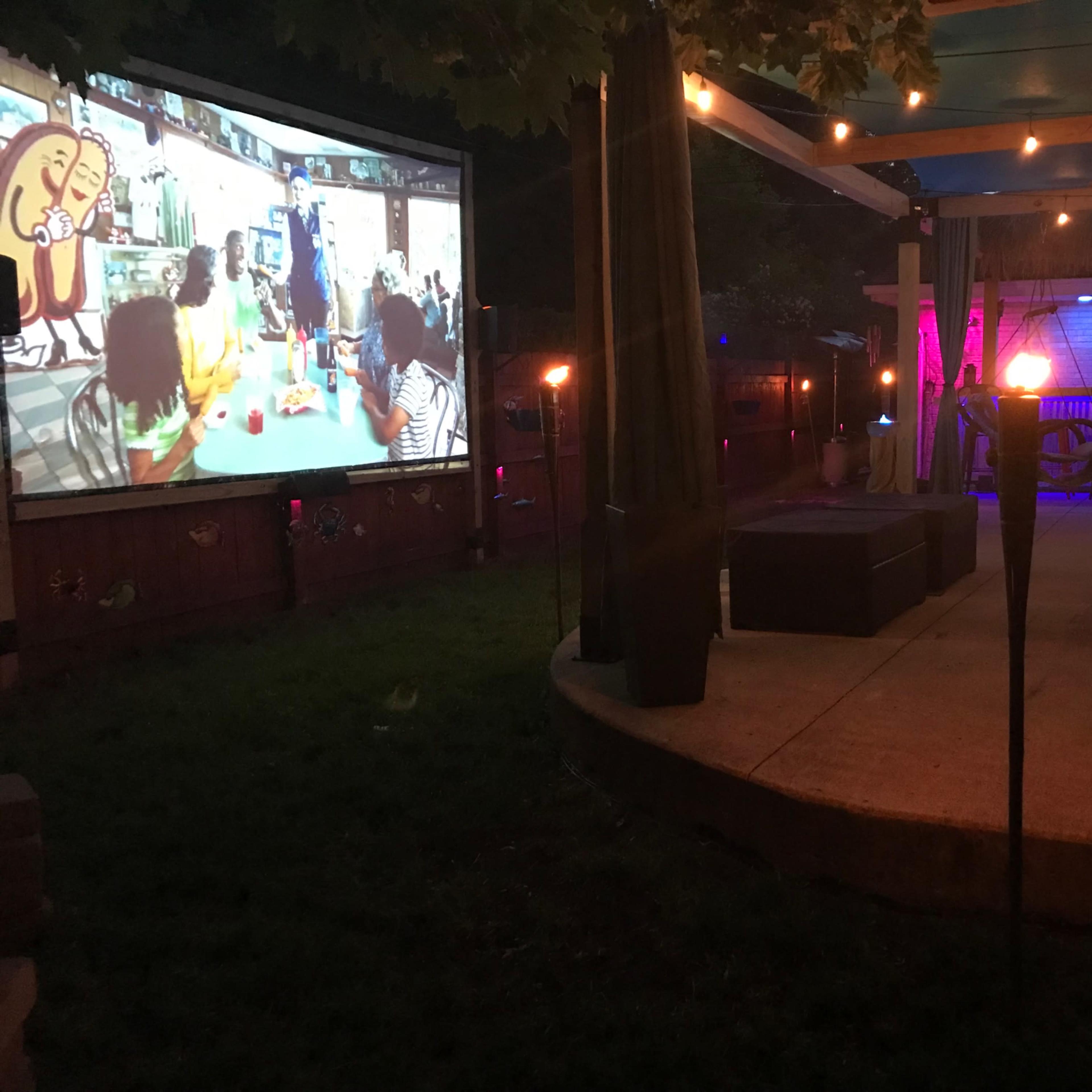 A large outdoor screen displays a colorful scene while a backyard area with seating and plants is illuminated by ambient lighting.
