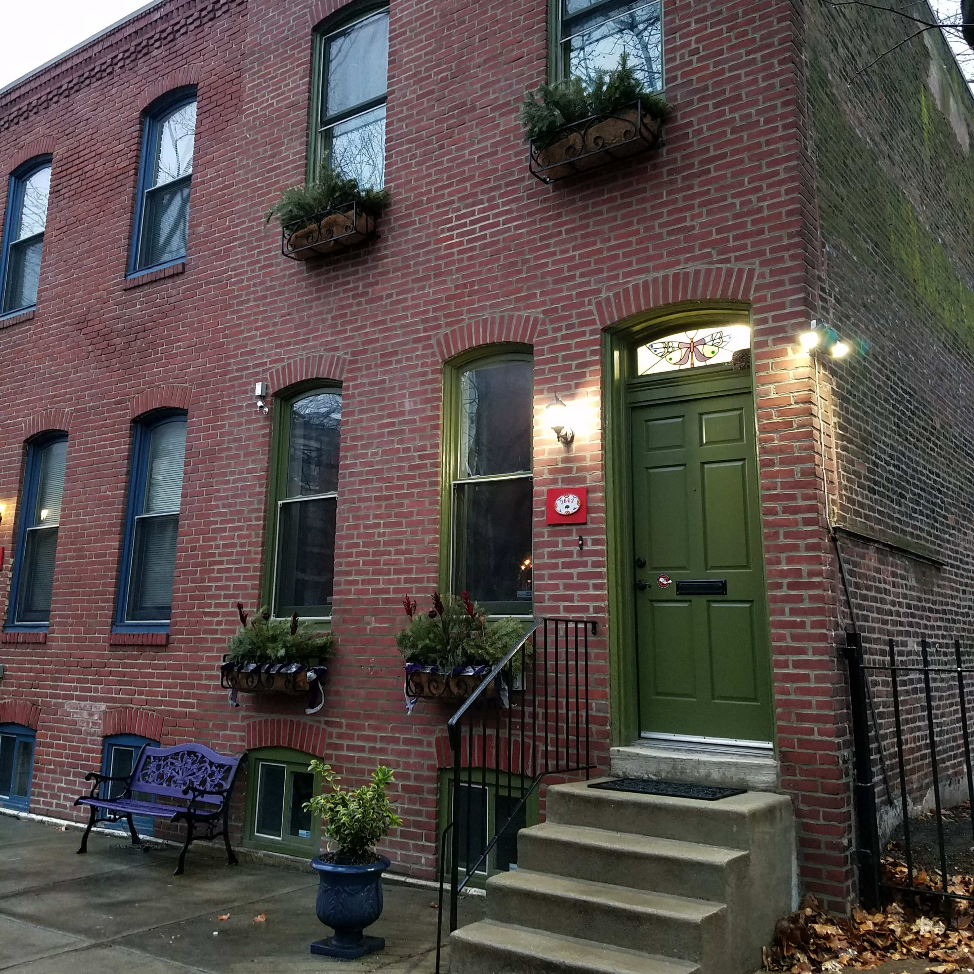 A brick townhouse with green door and window frames, featuring potted plants on the windowsills and a small staircase leading to the entrance.