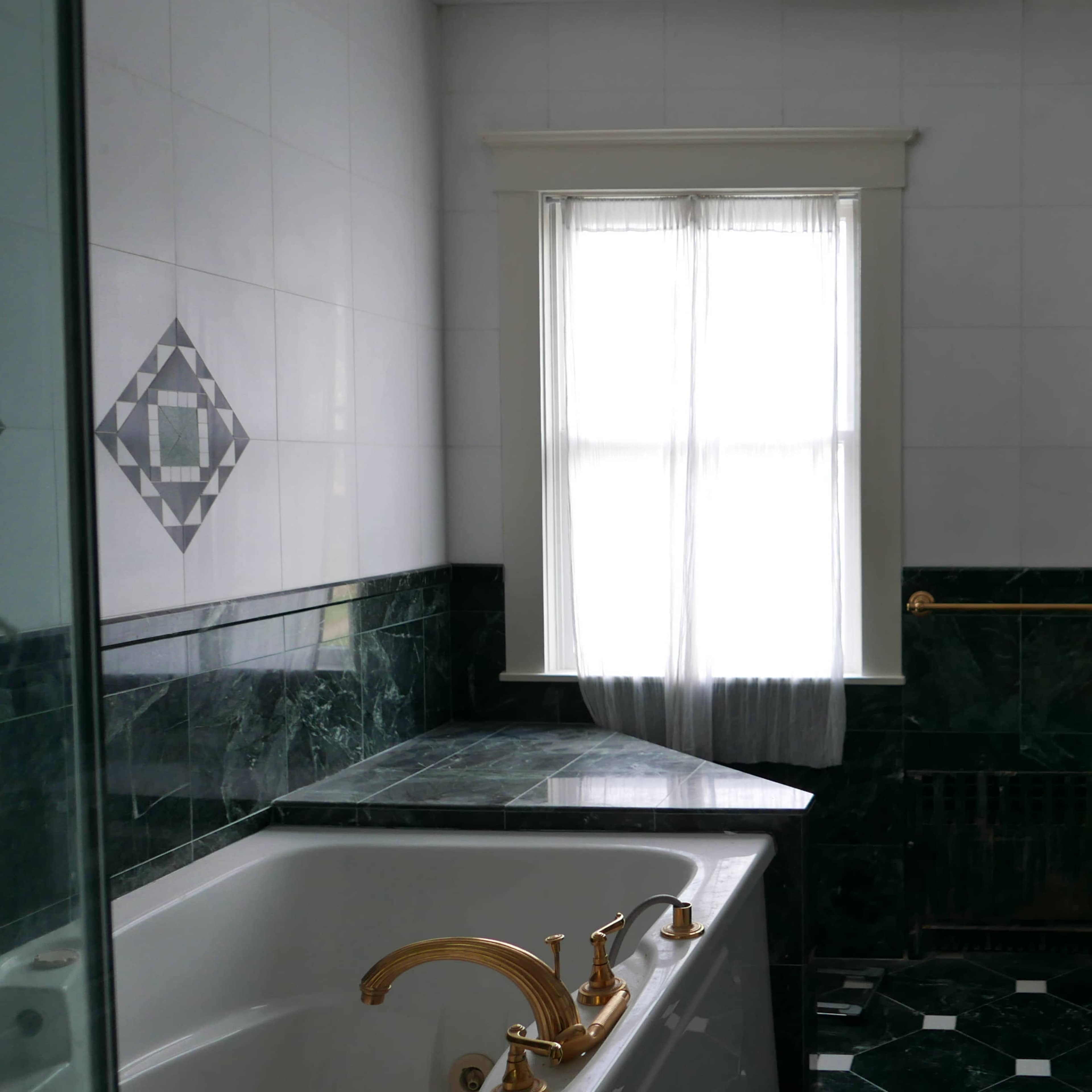 The bathroom features a marble bathtub, a window with sheer curtains, and dark green tile accents along the walls and floor.