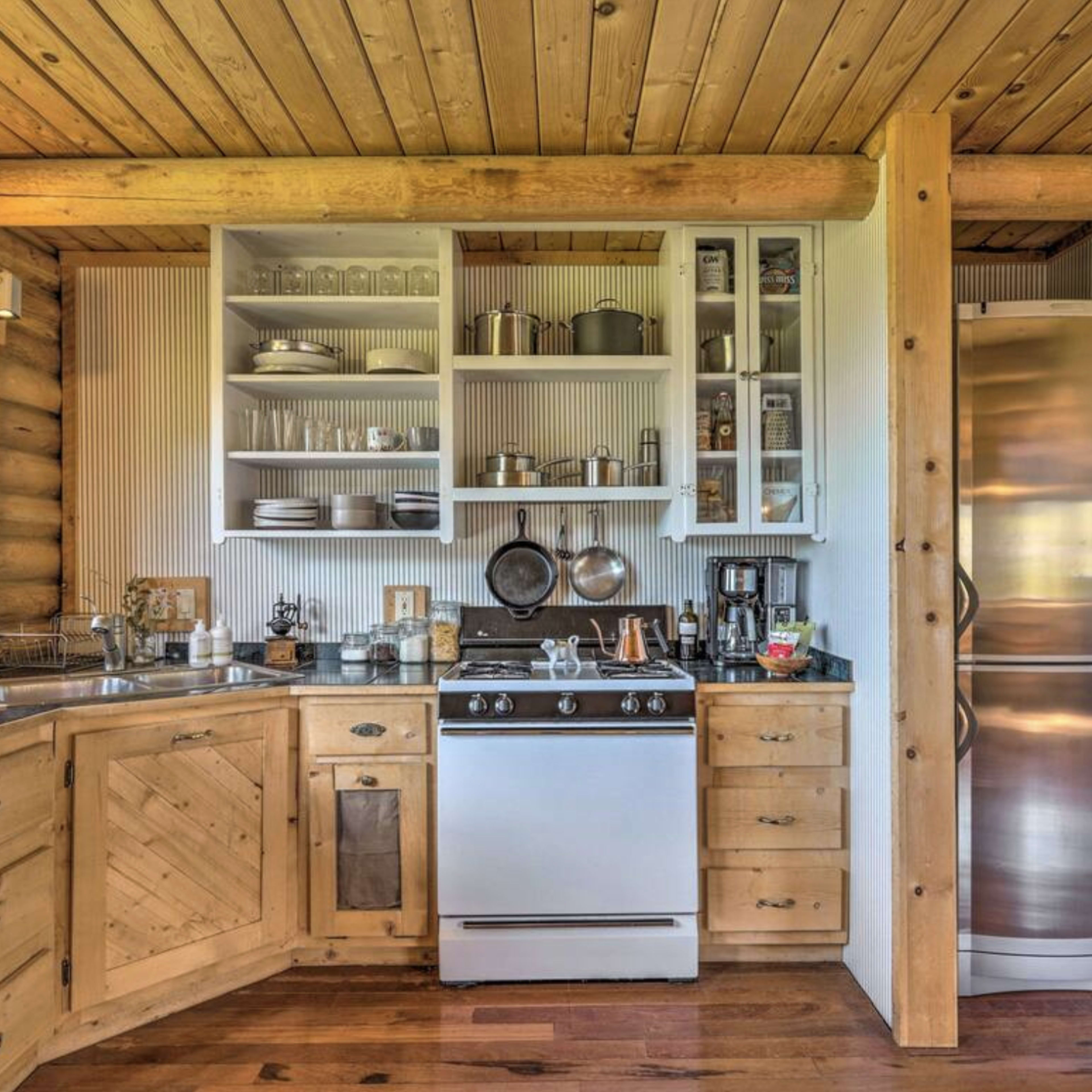 A rustic kitchen with wooden cabinets, a stainless steel refrigerator, a gas stove, and shelves displaying dishware and cooking utensils.