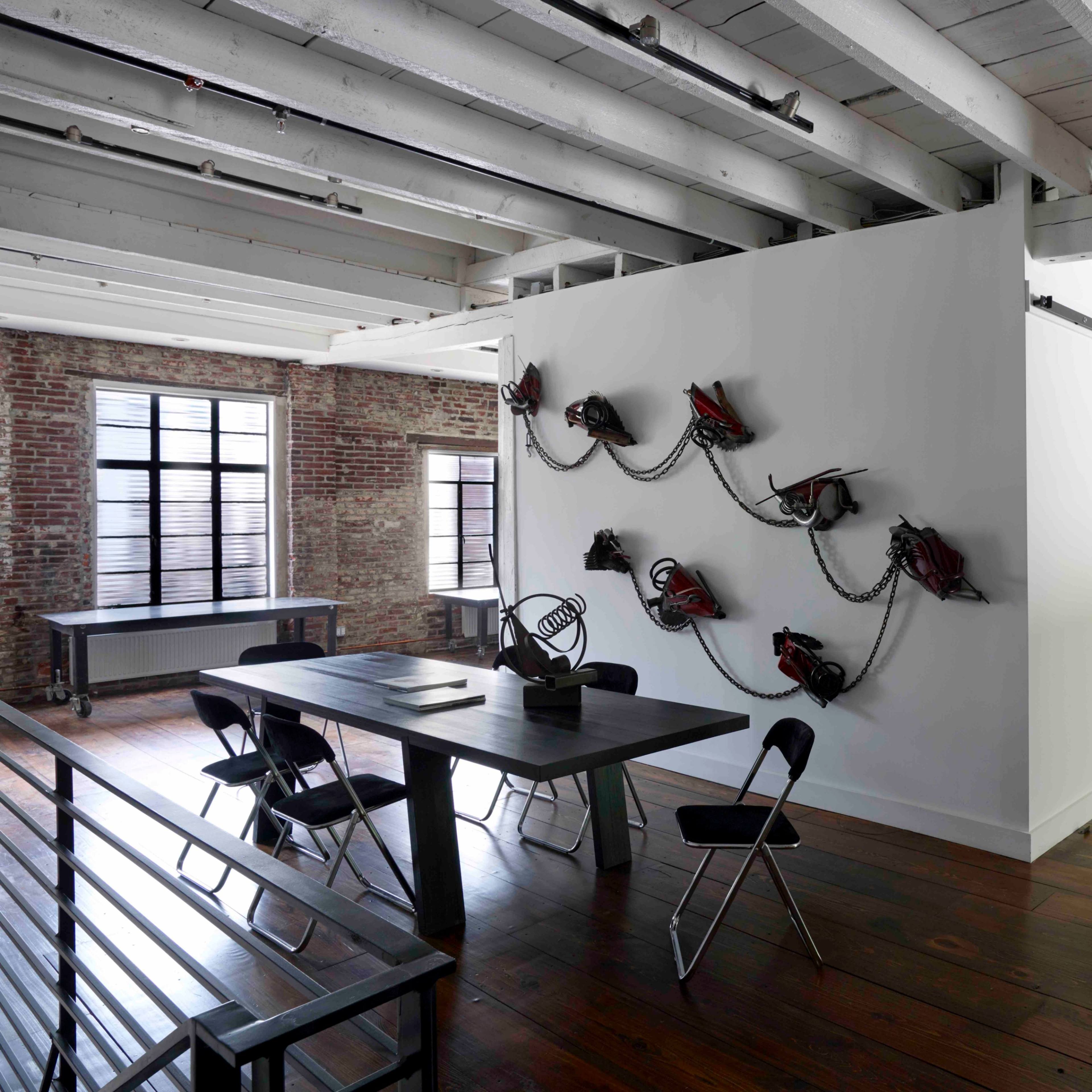 A modern room featuring a long table with chairs, a white wall adorned with chains and tools, and colorful abstract artwork on another wall, all set in a space with exposed brick and wooden beams.