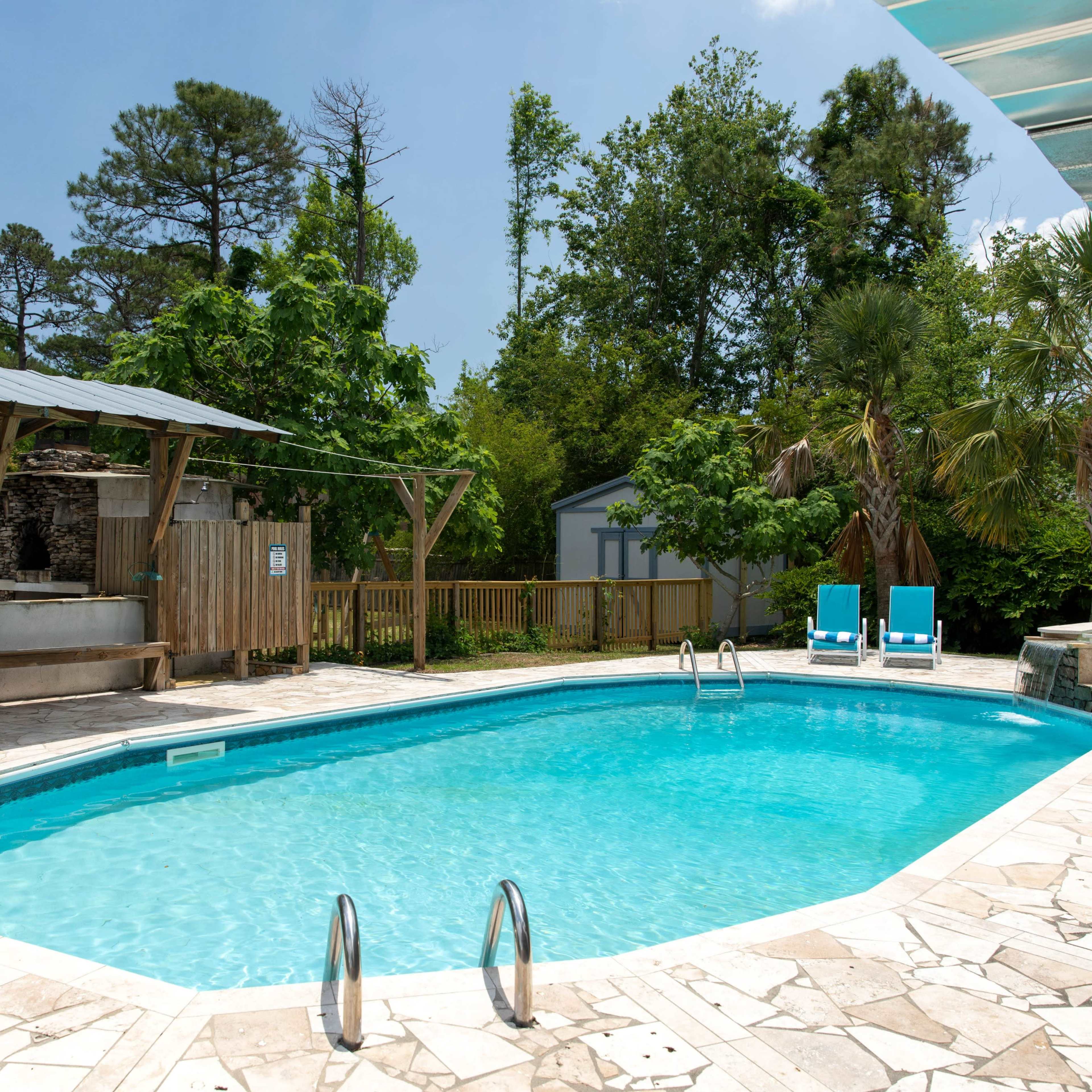 A rectangular swimming pool surrounded by stone decking, with lounge chairs on one side and a wooden gazebo in a lush, green backyard.