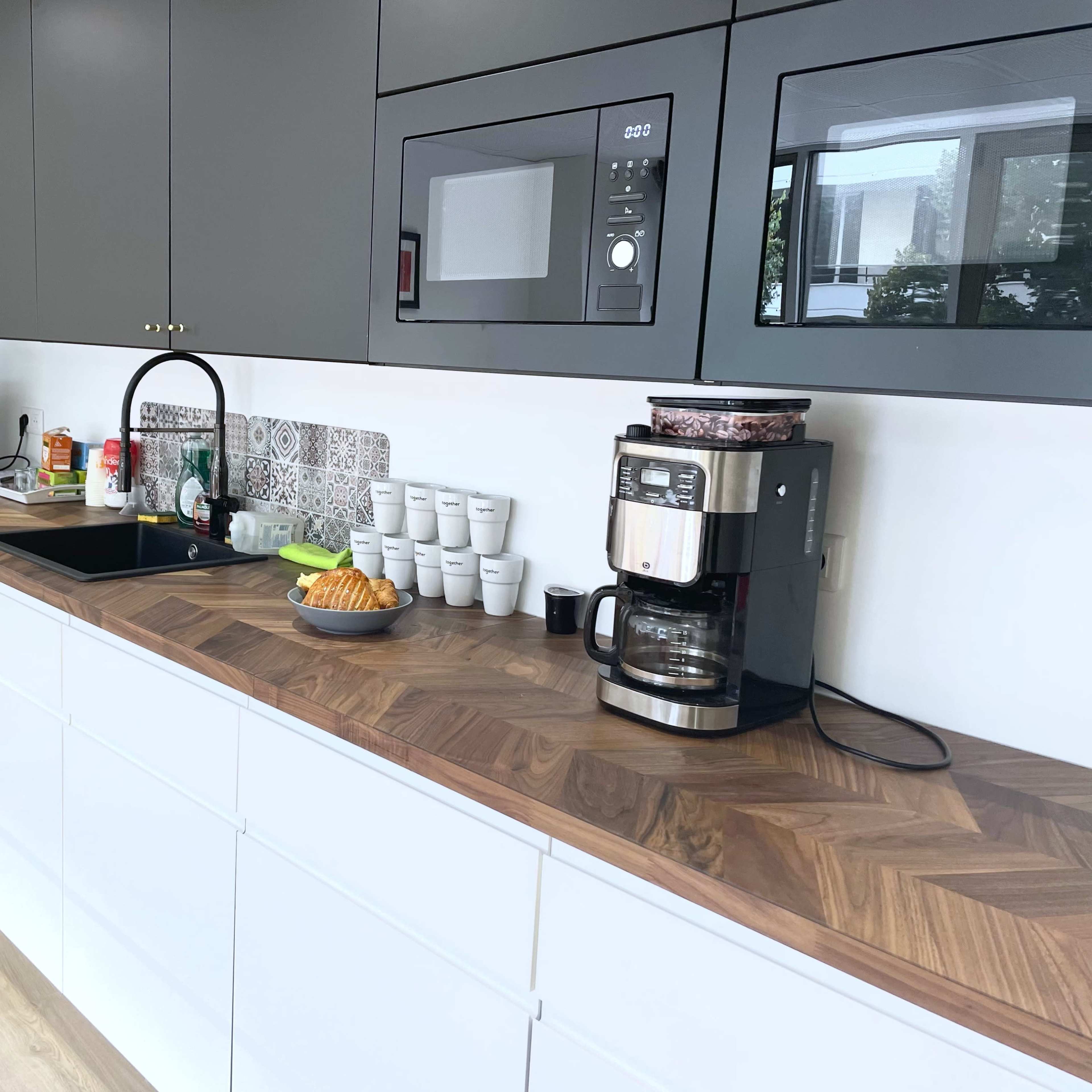 A kitchen countertop features a coffee maker, a sink, and various appliances, with several cups and a plate of pastries arranged neatly.