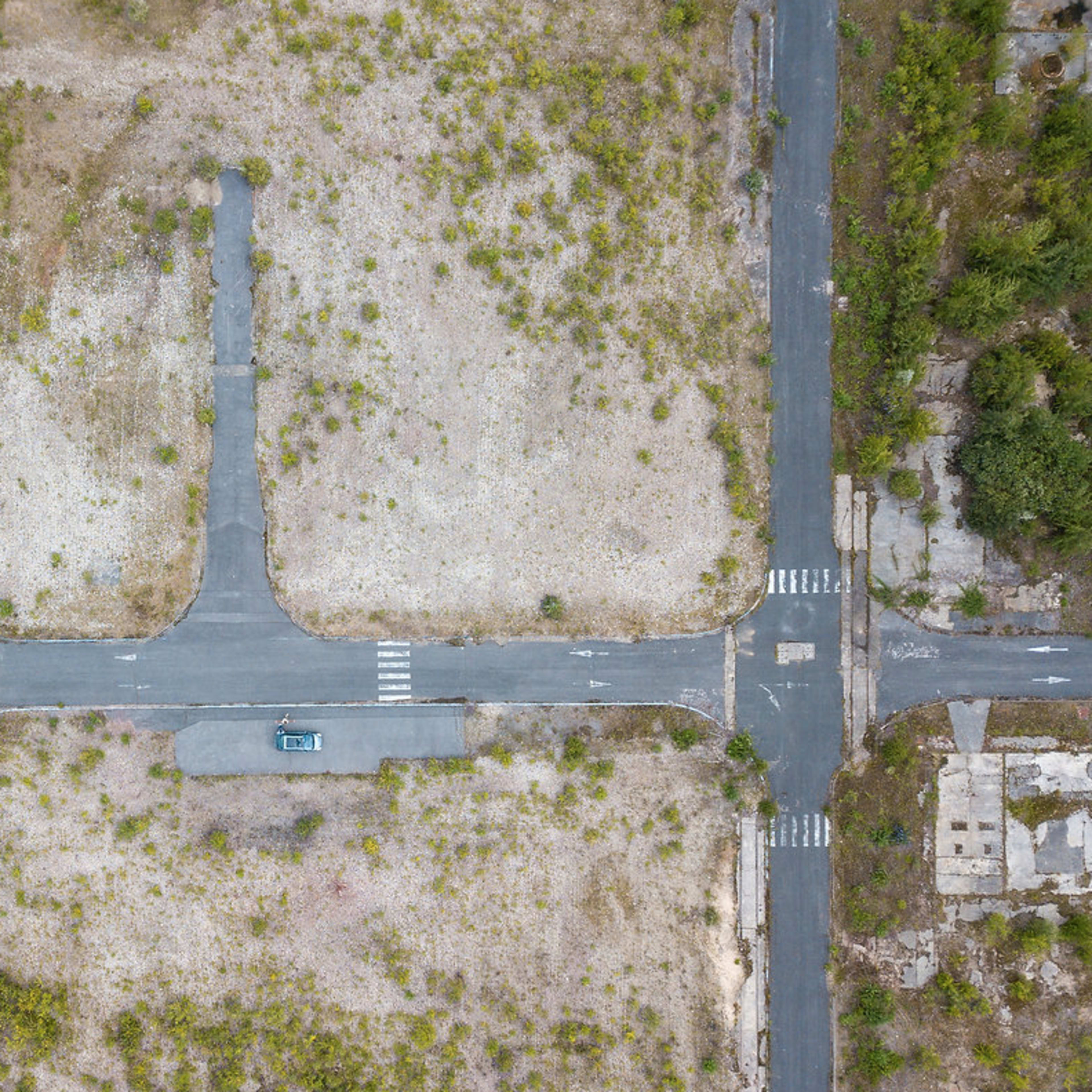 An aerial view shows a vacant intersection surrounded by overgrown vegetation and sparse asphalt roads.