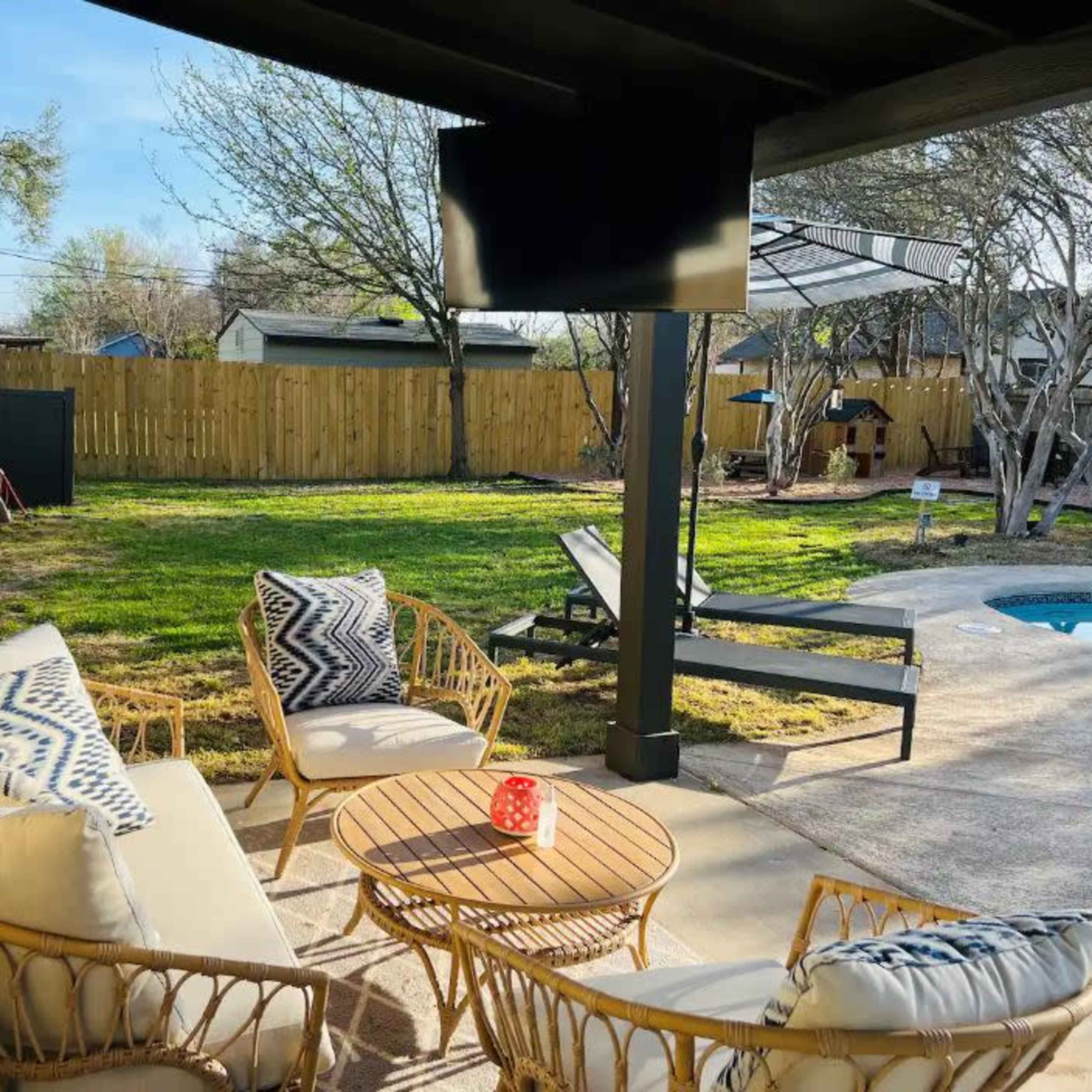 A patio area features a seating arrangement with wicker furniture, a small table, and a mounted television, overlooking a green backyard with trees and a pool.