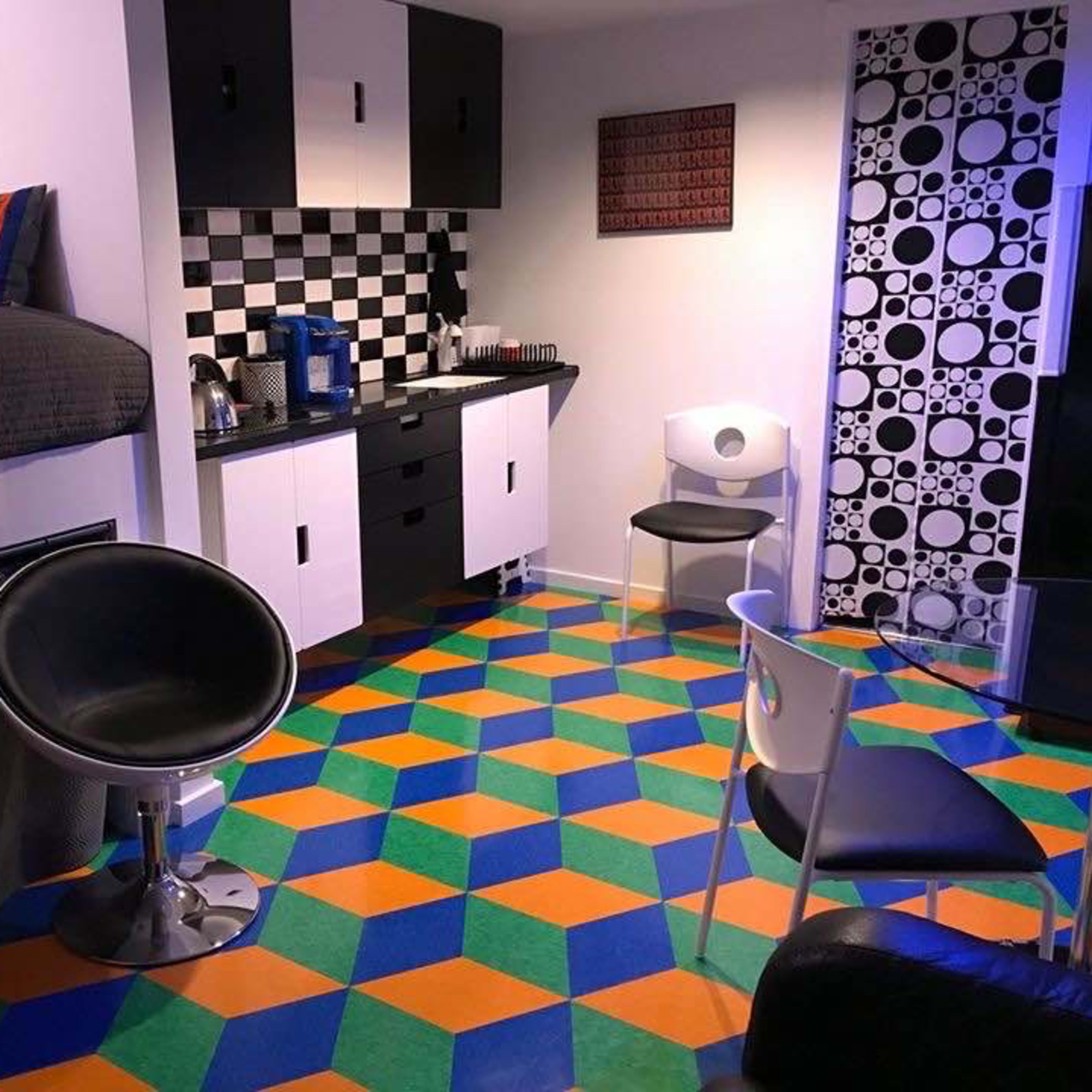 A small, modern room featuring a lofted bed, a checkered kitchen area, and a colorful geometric-patterned floor.