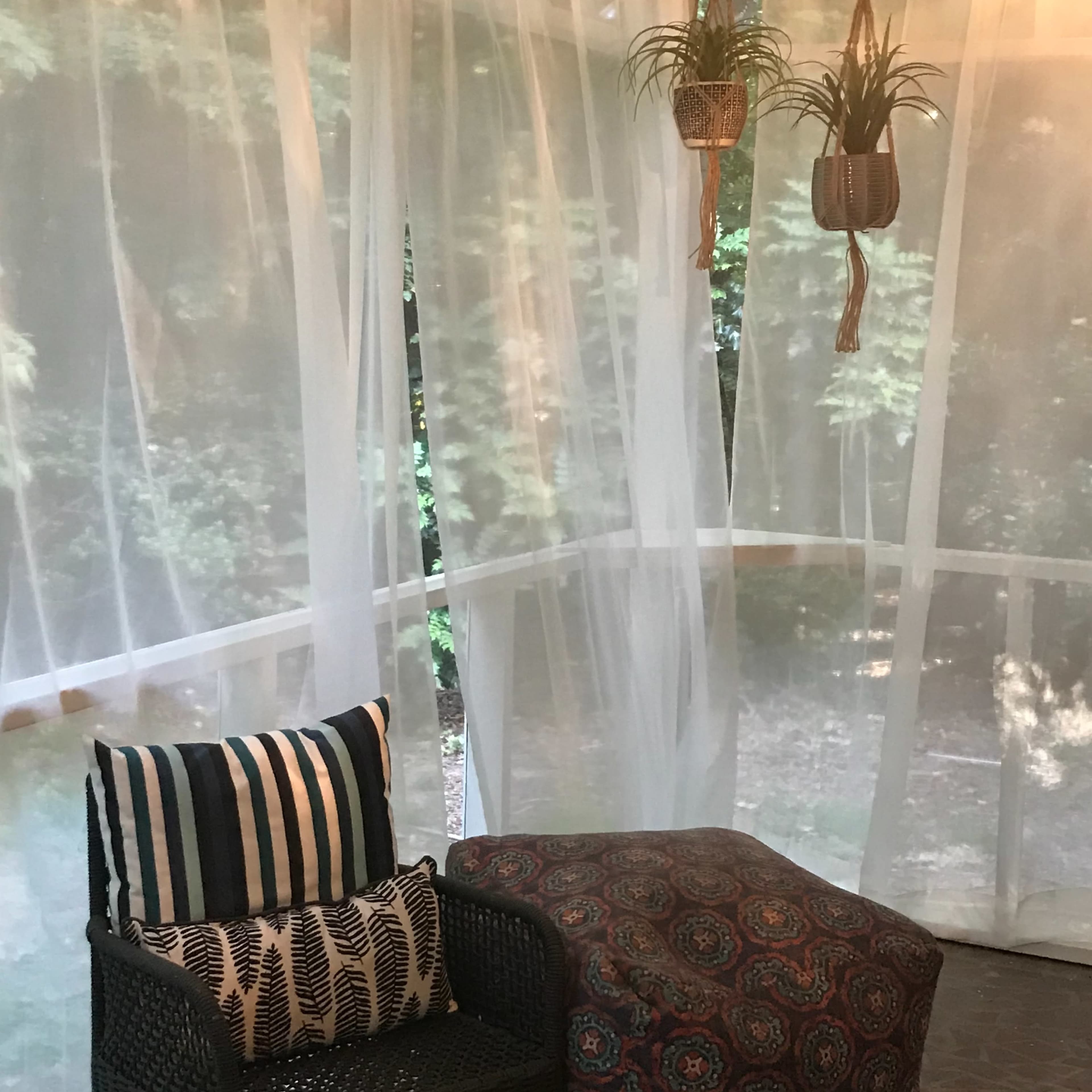 A screened-in porch with sheer white curtains, a hanging plant, string lights, and a chair with patterned cushions.