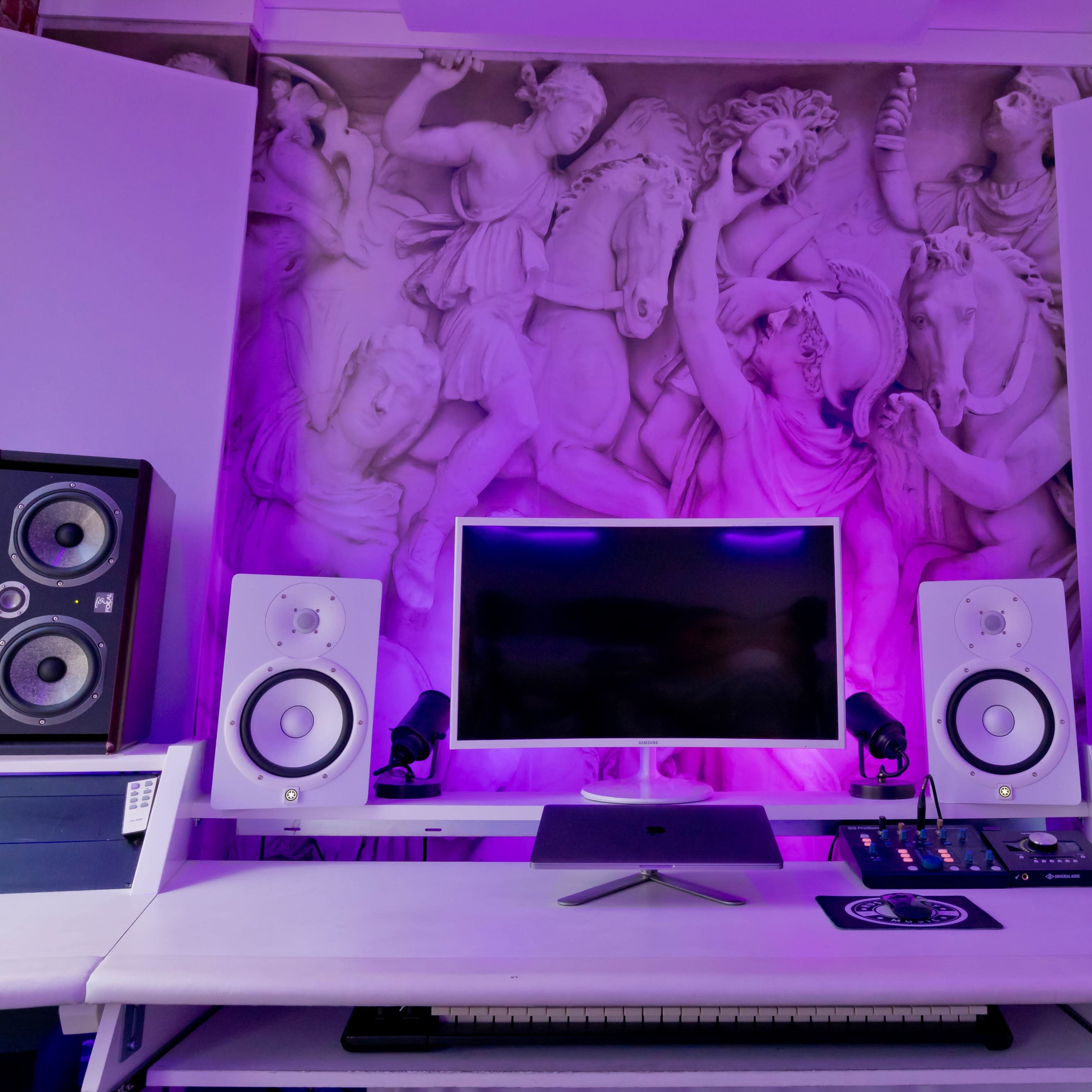 A music production setup features a central monitor flanked by studio speakers, with a decorative wall mural in the background.