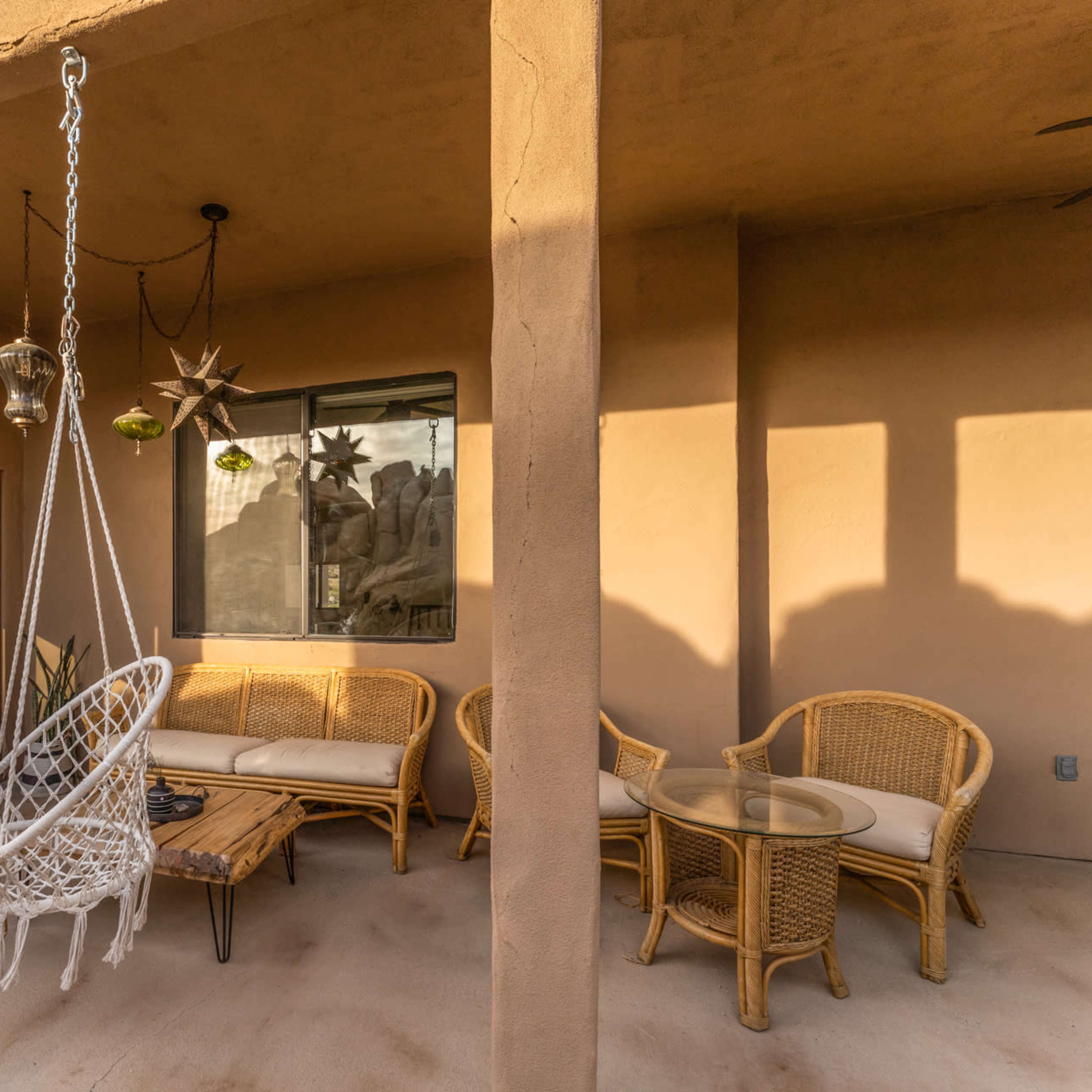 A patio with two hanging macramé chairs, a rattan sofa, a glass-top table, and a window with decorative items.