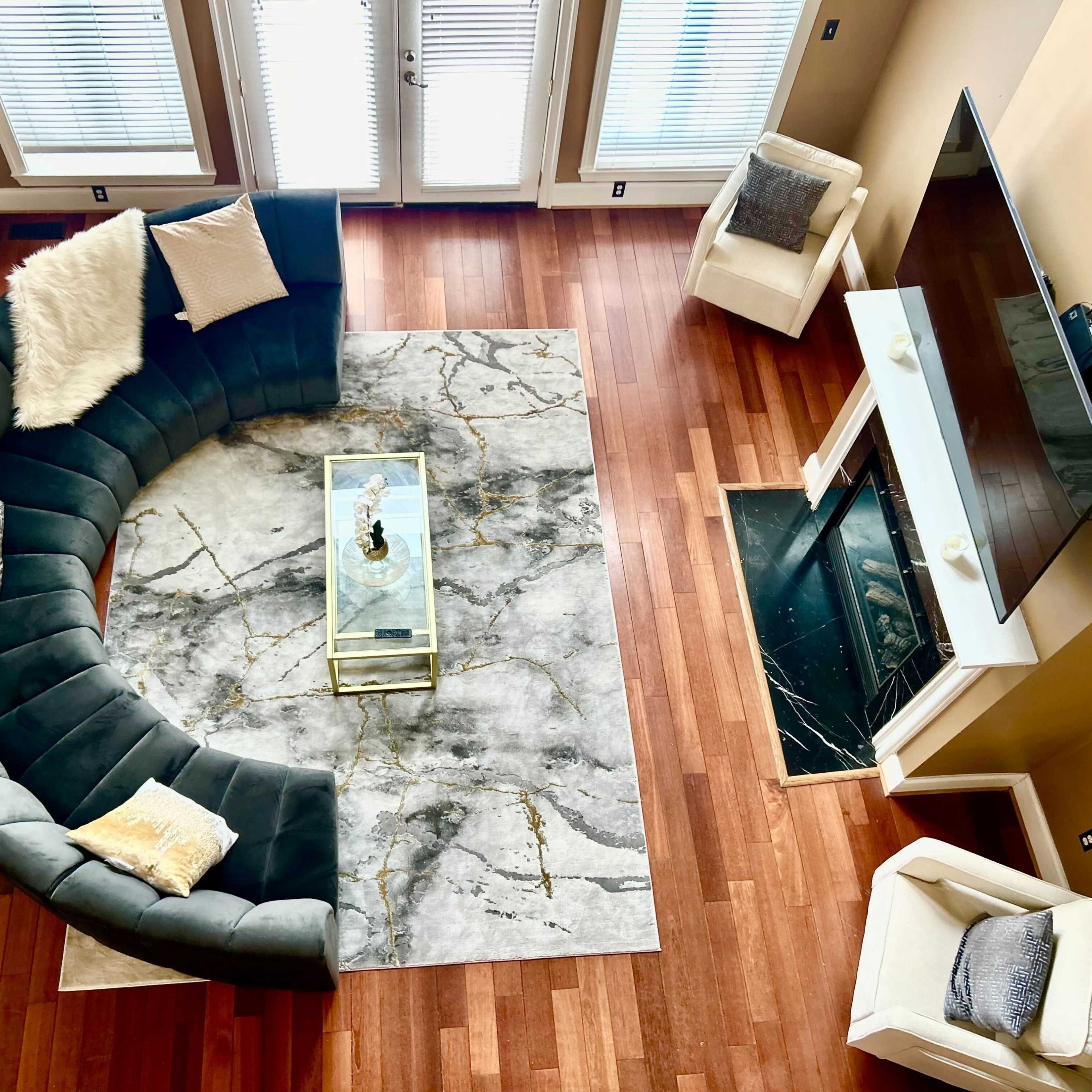 A modern living room features a curved gray sectional sofa, a marble-patterned rug, a glass coffee table, and a flat-screen TV above a fireplace.
