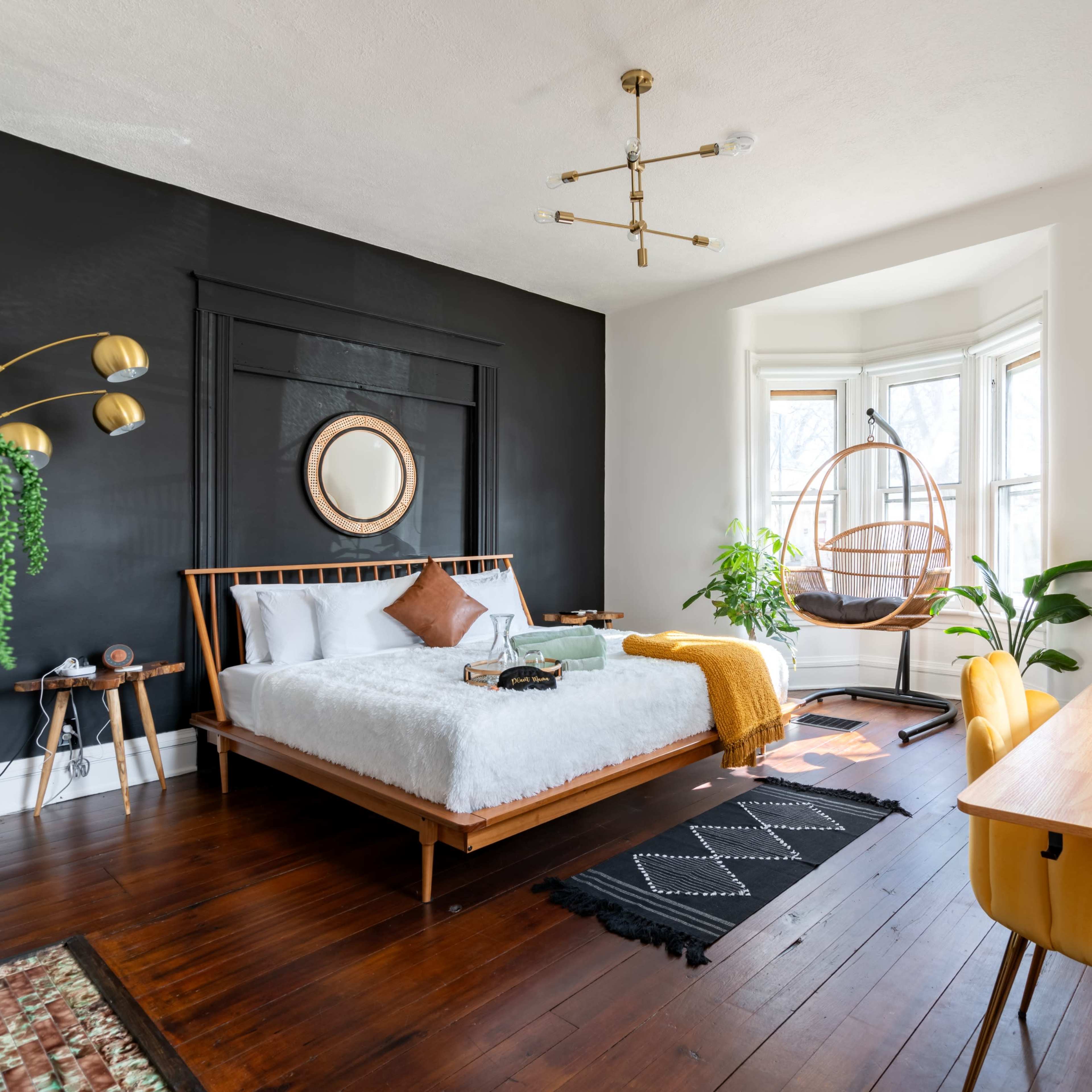 A modern bedroom featuring a white bed with decorative pillows, a hanging chair, a wooden desk with a yellow chair, and large windows allowing natural light.