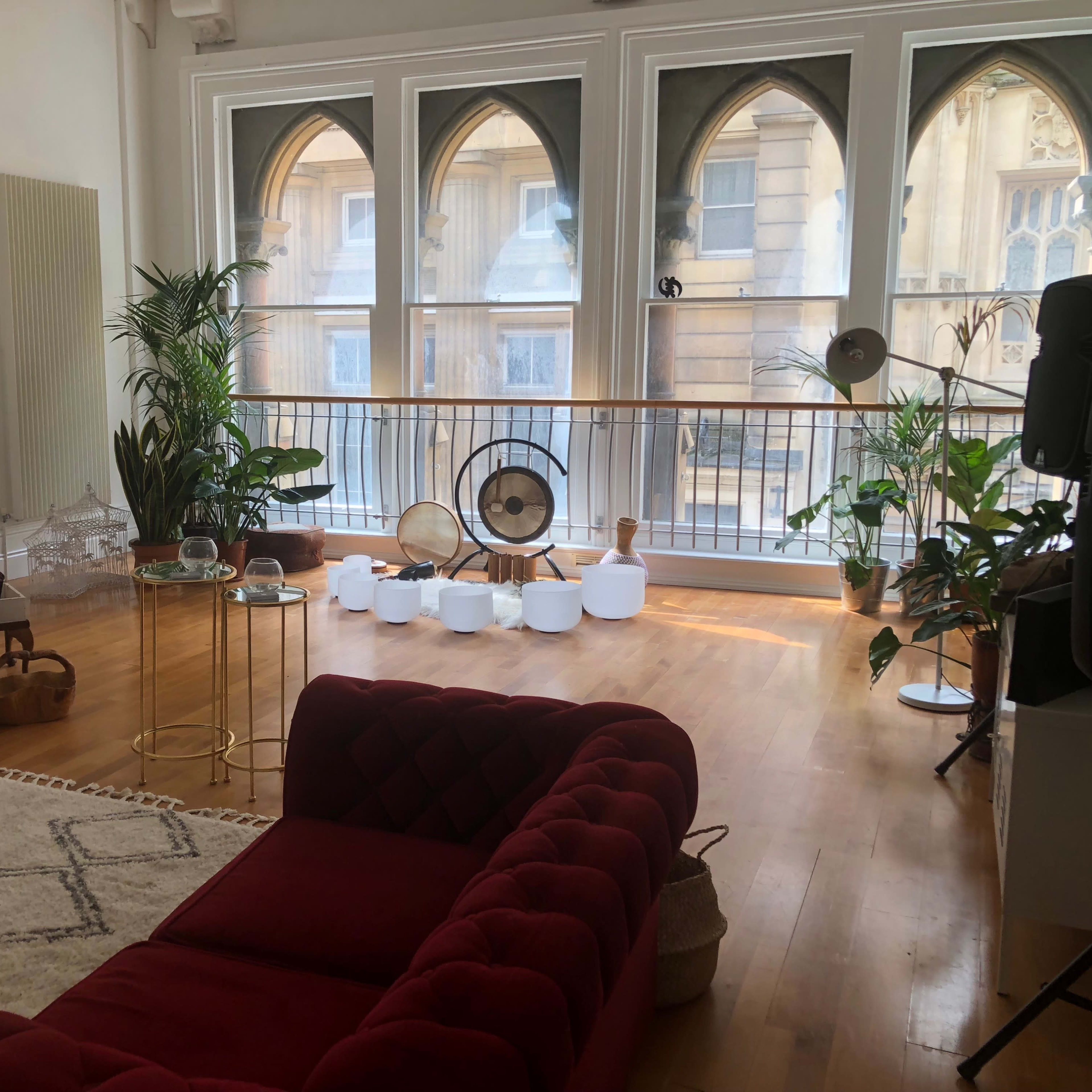 A modern living room with large arched windows, a mix of plants, a red sofa, and various decorative items.