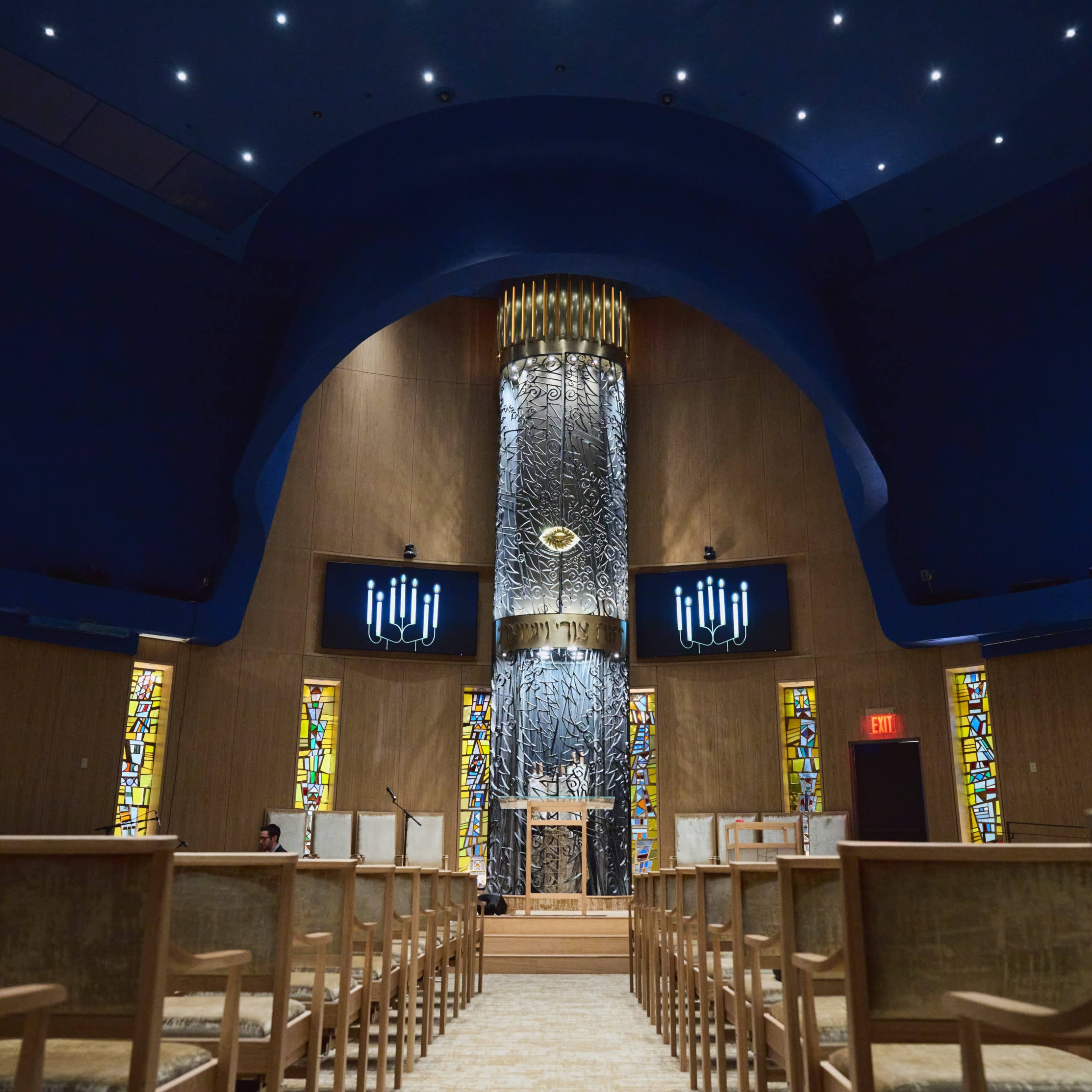 The interior of a synagogue features a central ark with a large stained glass window on either side and modern lighting illuminating the space.