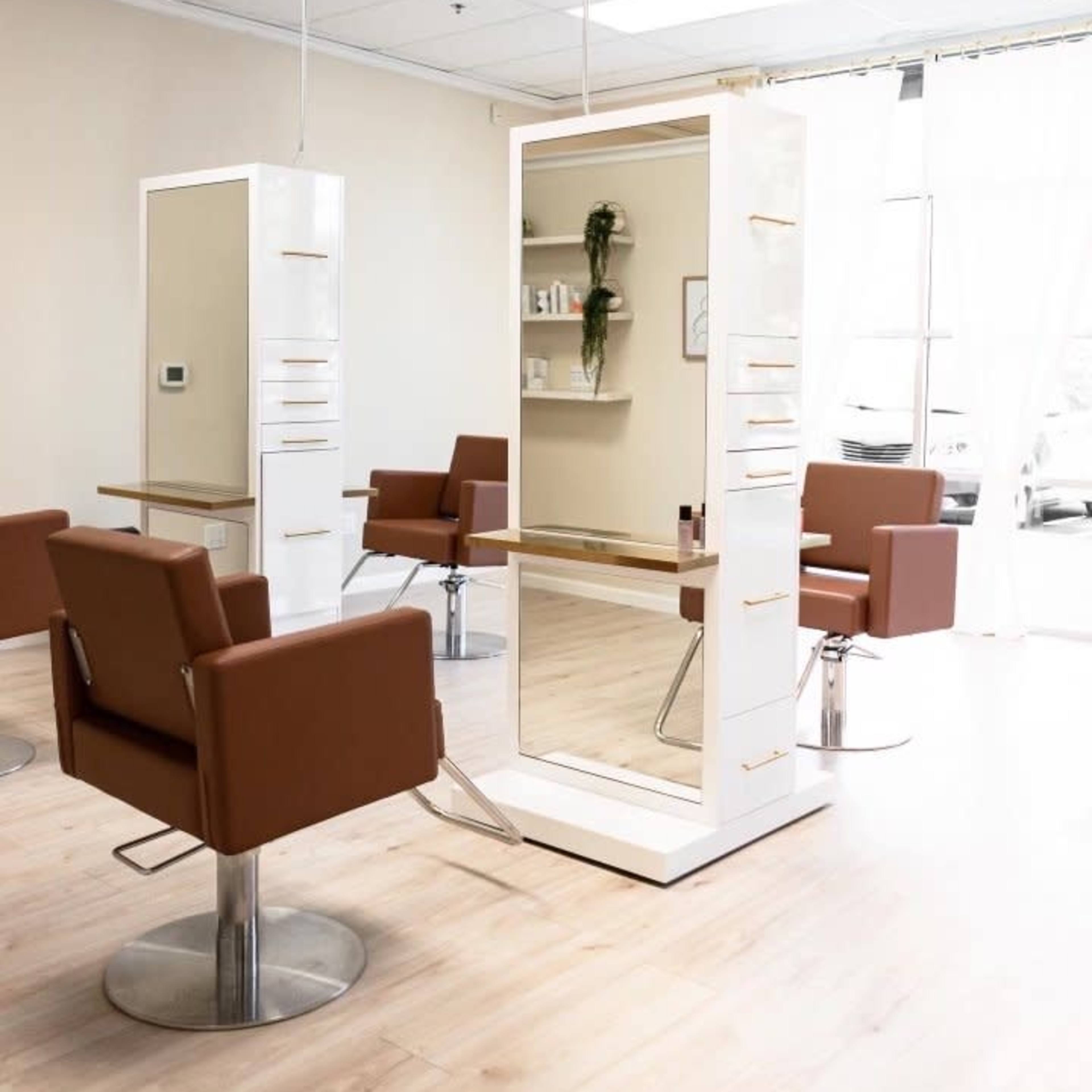 A modern hair salon with two styling stations featuring large mirrors and brown chairs arranged around a light-colored wooden floor.