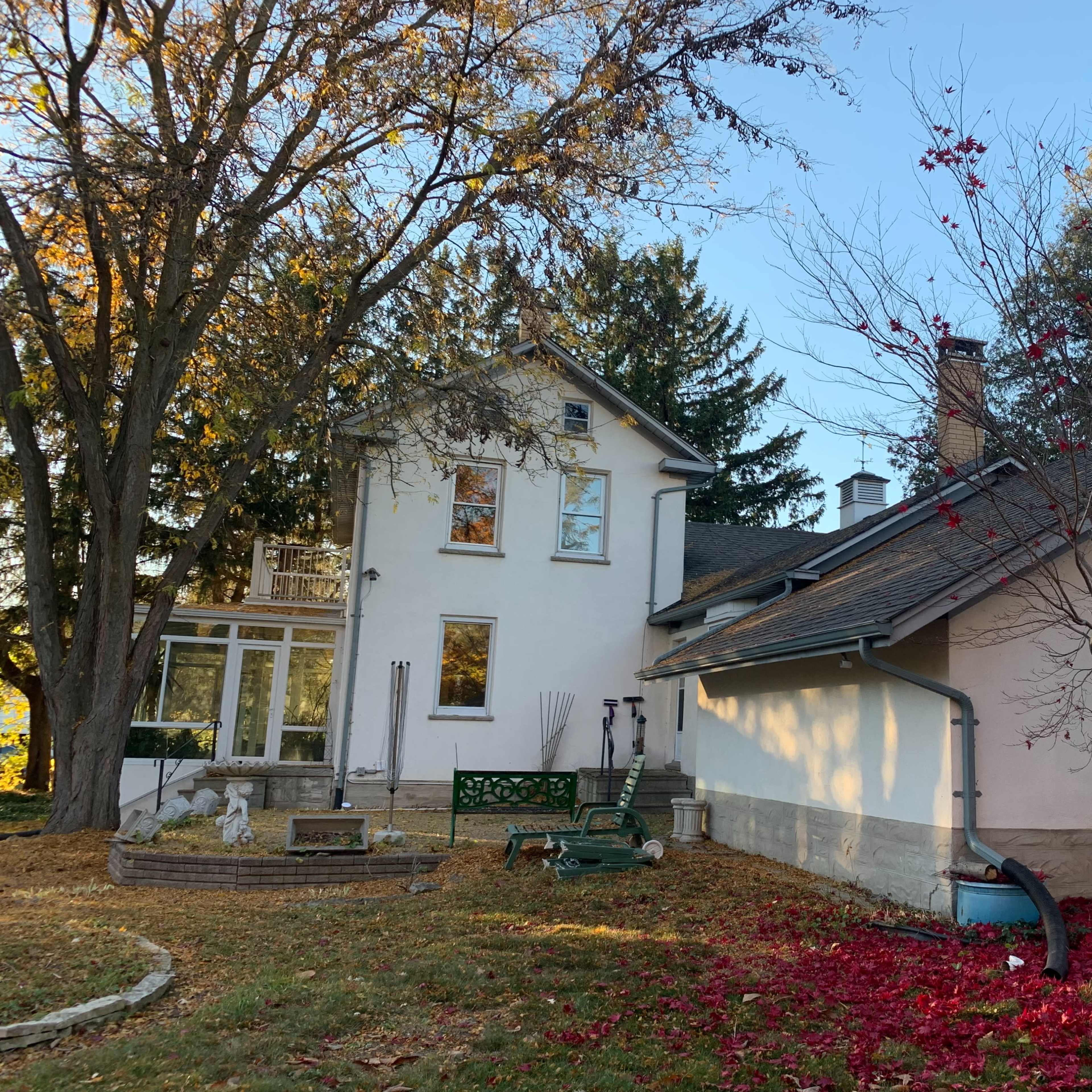 A two-story house with a green balcony and a side garden is surrounded by trees and decorative landscaping, featuring fallen red leaves.
