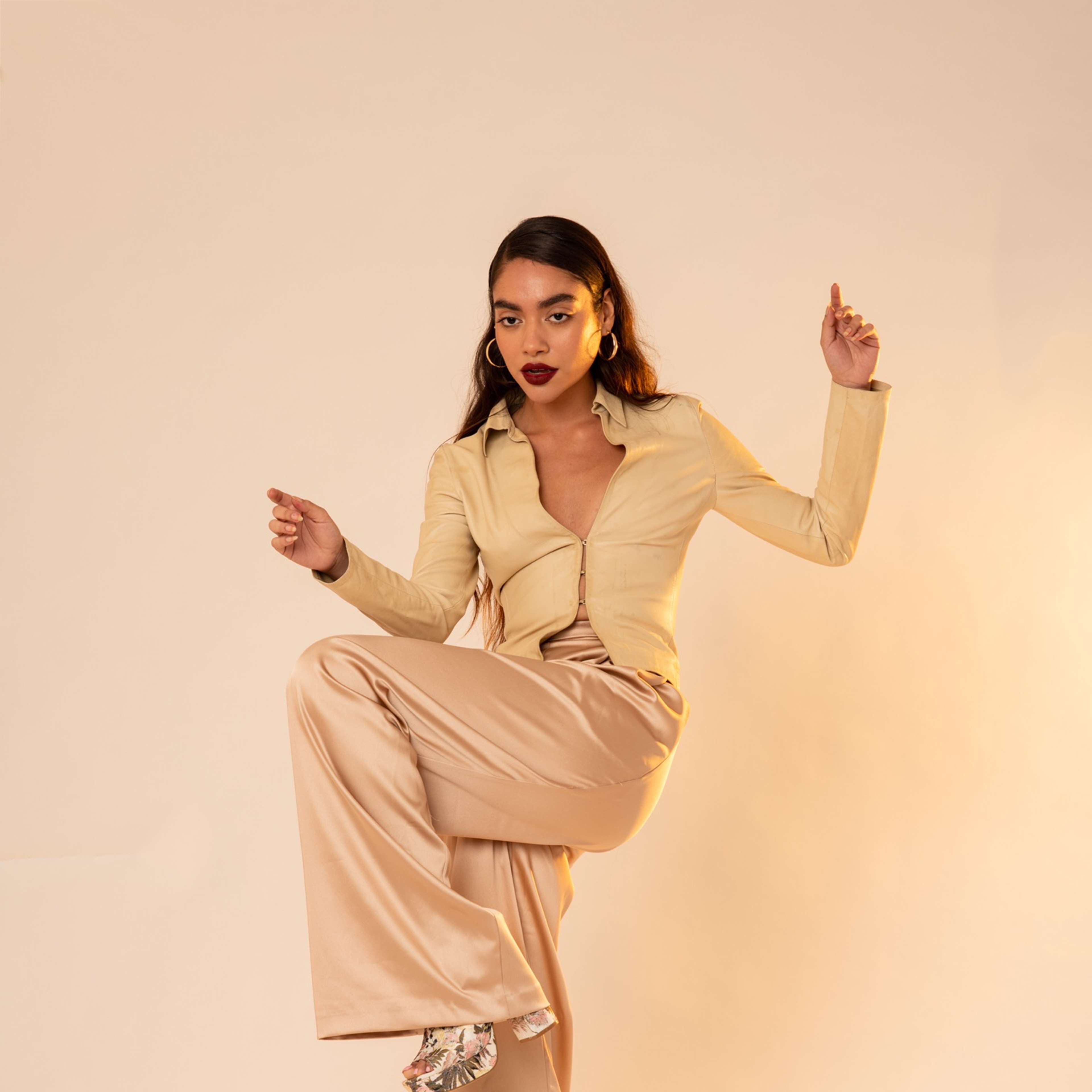 A model poses with one leg raised and arms outstretched, wearing a beige outfit and platform shoes against a light background.
